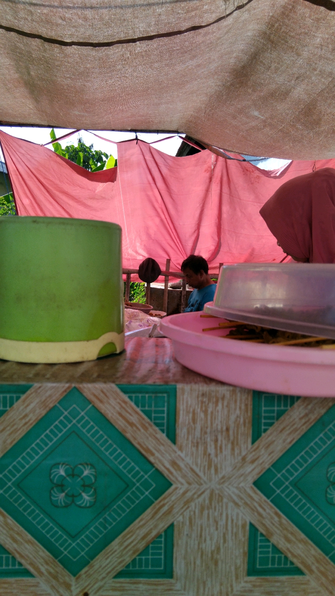 An overhead shot captures a rustic outdoor dining area, likely a roadside stall, in Batang, Indonesia. A pink tarp, possibly a makeshift awning, covers the scene, allowing glimpses of lush green foliage and blue sky beyond. In the foreground, a vibrant green mug rests on a table with a patterned surface featuring diamond shapes in teal and beige. To the right, a pink tray holds skewers of food, partially covered by a clear plastic lid. In the background, a man in a blue shirt is seated at a table, facing away from the camera. The overall ambiance suggests a simple, everyday meal being enjoyed outdoors.