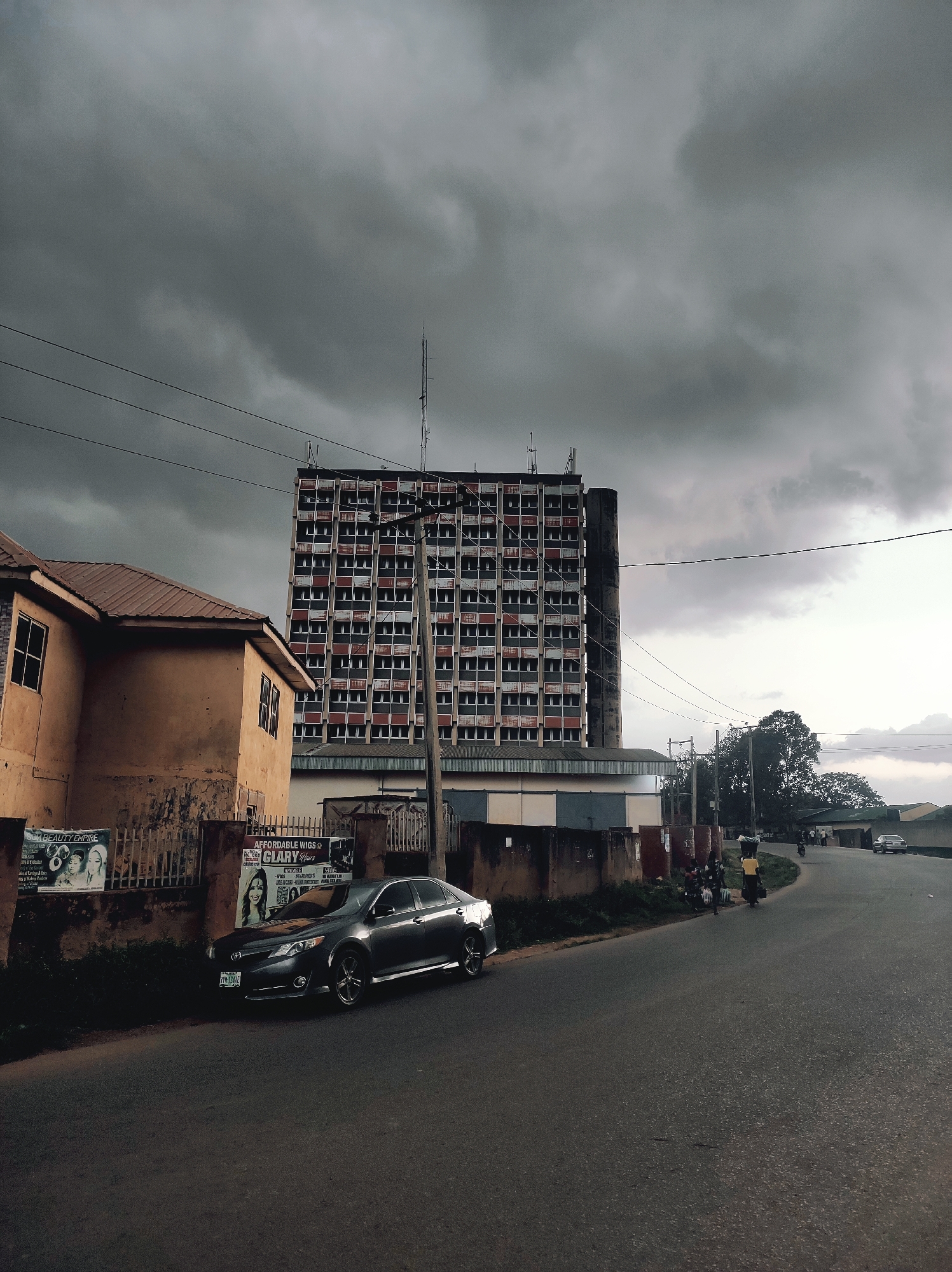 The image depicts a street scene in Jos, Nigeria, under a heavily overcast sky. A tall, multi-story building with a grid-like facade dominates the background. The building has multiple floors with visible windows. In the foreground, a residential building stands to the left, next to the street. A dark-colored sedan is parked along the side of the road, near a fence covered with posters. Further down the road, there are individuals walking with items carried on their head. A couple of vehicles are also visible.
