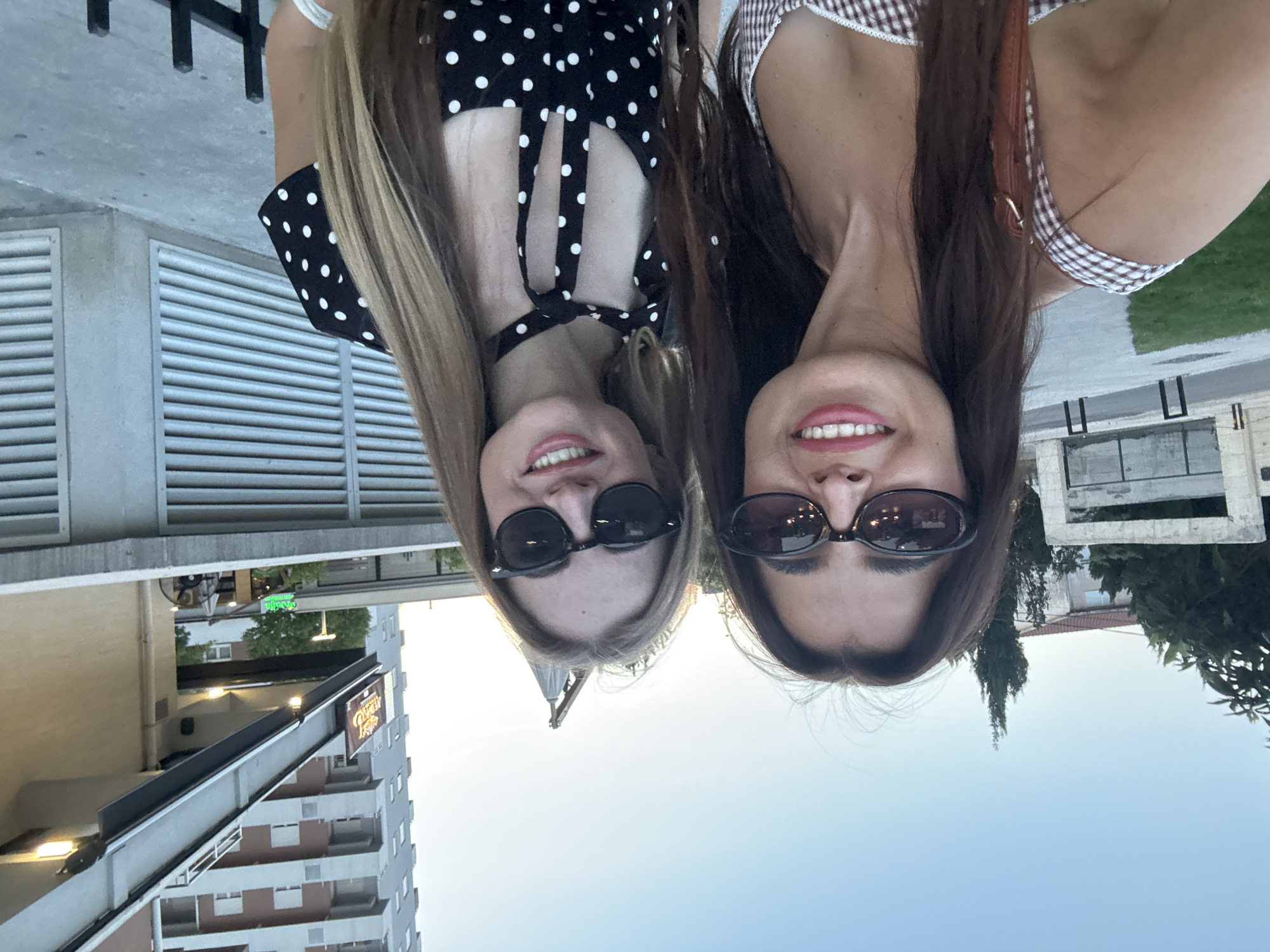 Two women are posing for a selfie outdoors in front of a building with a light-colored facade and a tall, multi-story building in the background. The woman on the left has long, dark hair and is wearing a brown and white checkered top and sunglasses. The woman on the right has long, blonde hair and is wearing a black and white polka dot dress and sunglasses. Both women are smiling and appear to be in good spirits. The background includes some trees, a street, and a fence. The lighting suggests it is either early morning or late afternoon.