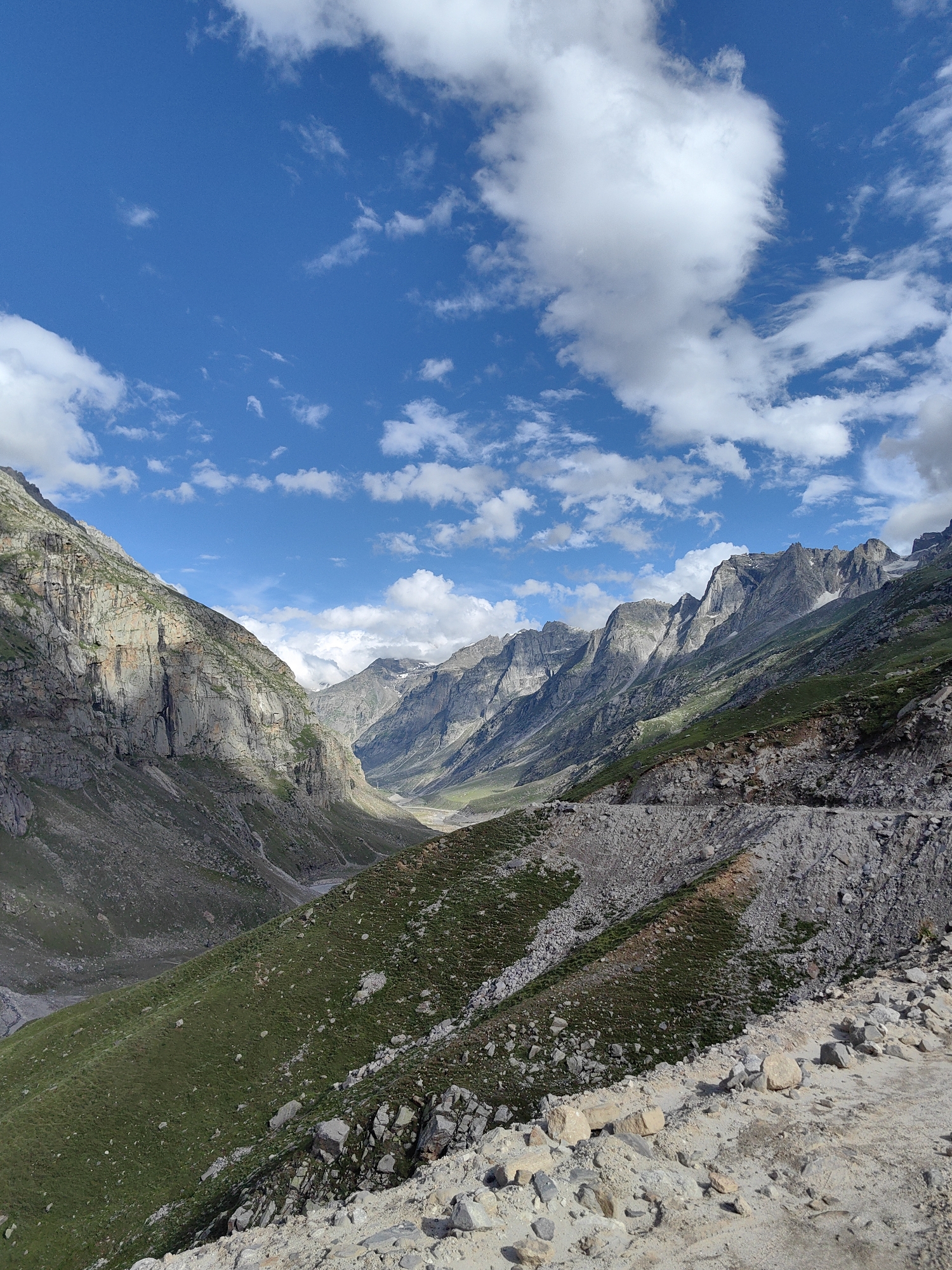 The image depicts a vast mountainous landscape under a bright blue sky dotted with fluffy white clouds. The terrain is characterized by steep, rocky slopes covered in patches of green grass. In the foreground, a dirt and gravel path winds its way along the mountainside. The middle ground shows a valley with a riverbed and more grassy slopes leading up to towering, rugged mountains that stretch into the distance. The lighting suggests it is daytime, with sunlight casting shadows across the mountains, highlighting their texture and depth. There are no people or man-made structures visible in the scene, emphasizing the natural and remote environment. The location is identified as being in the vicinity of the city of Bashisht, India, a region known for its Himalayan scenery.