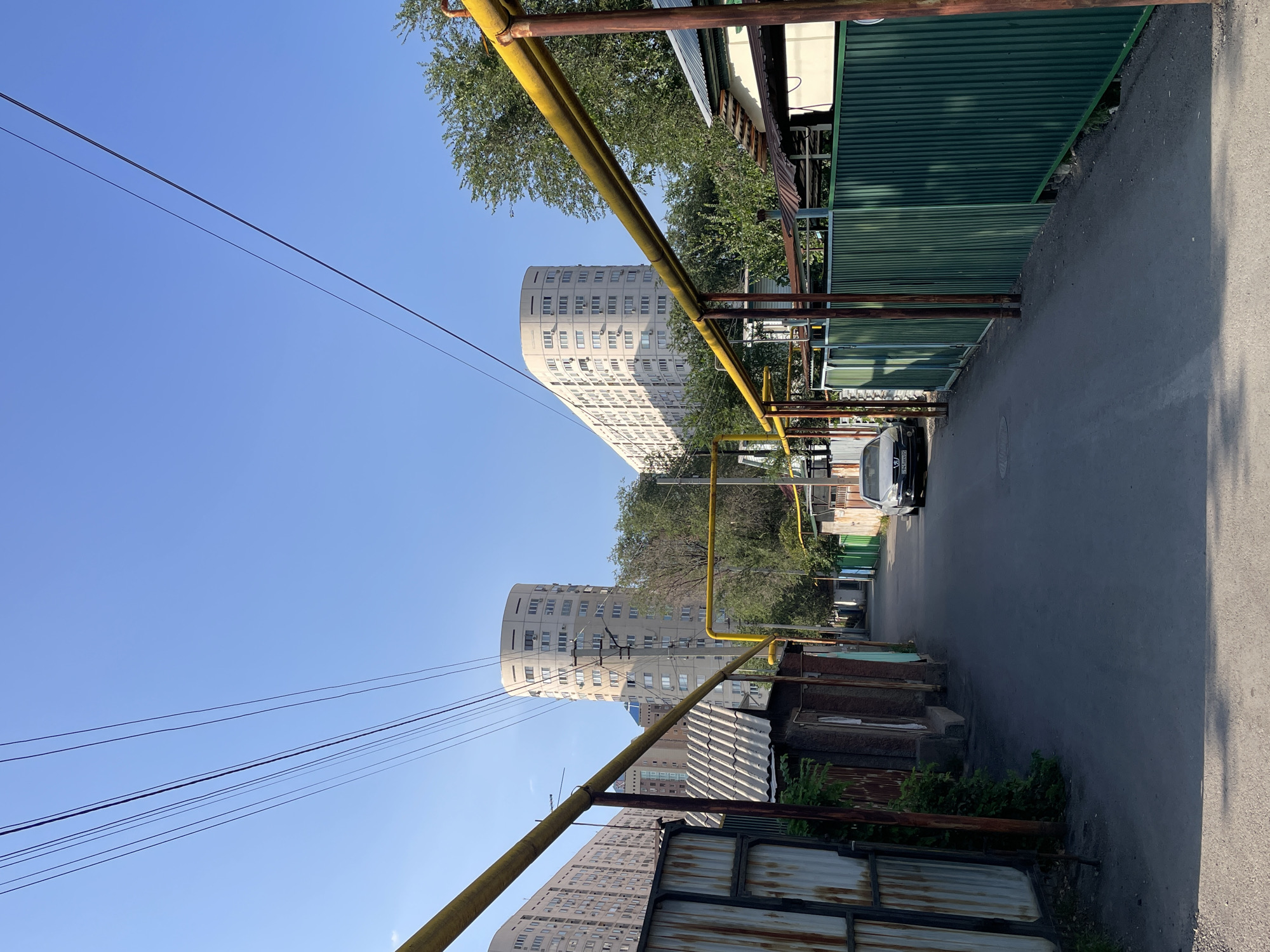 An outdoor, eye-level, wide-angle shot captures a street in Almaty, Kazakhstan. The scene is dominated by utility poles and wires crisscrossing the clear blue sky, casting shadows on the asphalt road. The street is lined with low-lying, weathered structures on the left and a corrugated metal fence on the right. In the distance, two distinctive, curved apartment buildings rise above the smaller structures, their beige facades punctuated by numerous windows. A silver sedan is parked on the right side of the street, its license plate visible. Lush green trees are interspersed between the buildings, adding a touch of nature to the urban environment. The overall impression is one of a residential street with a mix of older and newer architecture.