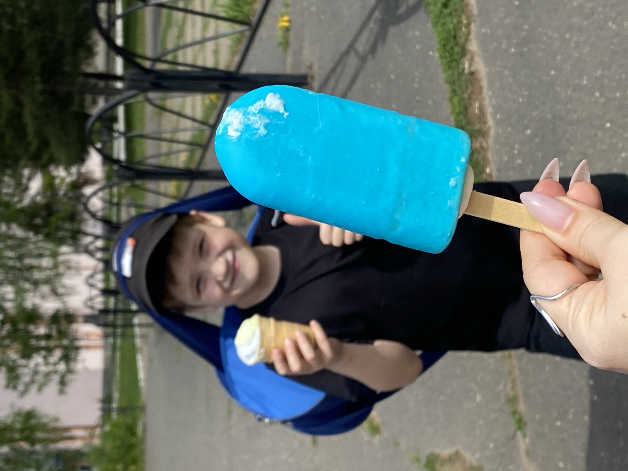 A sunny outdoor scene in what appears to be a park or recreational area. A child, possibly a young boy, is holding an ice cream cone in one hand. A person's hand is holding a large, bright blue popsicle. The child is slightly out of focus behind the popsicle, and appears to be smiling. The child is wearing a black shirt, a black baseball cap, and what may be a blue backpack. In the background is a black metal fence, trees, and part of a building. The ground is paved. There is a ring on one of the fingers of the hand holding the popsicle. Based on visual clues, it is possible that this image was taken in the city of Chaikovsky, Russia.