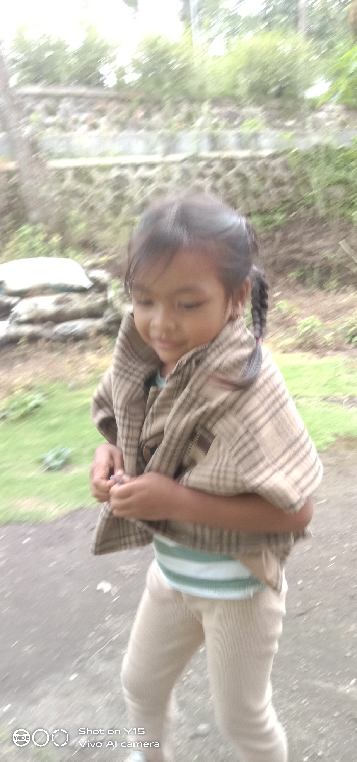 A young child with dark hair in two braids stands outdoors in Ranugedang, Indonesia. The child is wearing a light-colored striped shirt, light-colored pants, and a beige and brown plaid shawl draped over their shoulders. The child looks downwards and to the left, with hands clasped at chest height, seemingly holding a small, indistinct object. The immediate ground is a dark path, flanked by green grass. In the background, a multi-tiered stone or concrete retaining wall is partially visible, with blurred green foliage and trees beyond. The entire image exhibits significant motion blur and is out of focus.
