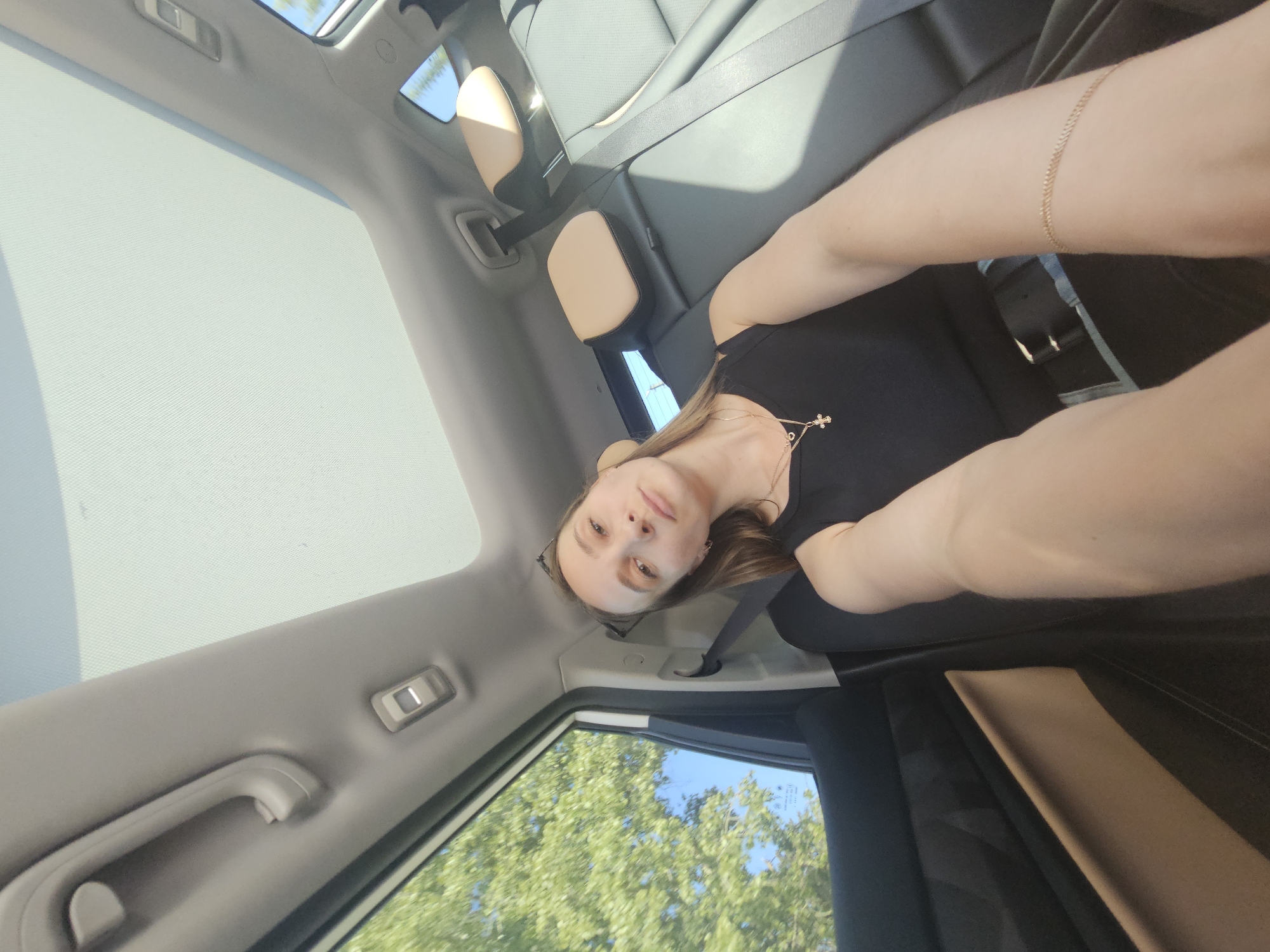 A woman is in the back seat of a car, possibly a ride share, facing towards the camera. The view outside the window shows lush green trees. The interior of the car has a sunroof. The woman has on a black top and gold jewelry. The location is uncertain, although the prompt indicates it may be Chaikovsky, Russia. There are no clear indicators in the image that confirm the location.