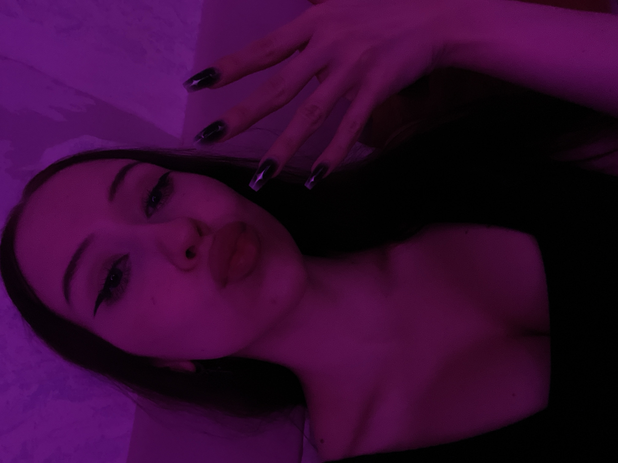 The image shows a close-up, selfie-style shot of a young woman with long, dark hair, wearing a dark top. Her face is tilted towards the camera, with her lips pursed. She is wearing dramatic winged eyeliner. Her right hand is raised, with long, dark, pointed fingernails. The lighting is predominantly purple, casting a strong magenta hue over the scene. The background appears to be an interior setting, with a textured wall visible. There is no indication of the city of Votkinsk, Russia, within the image itself.