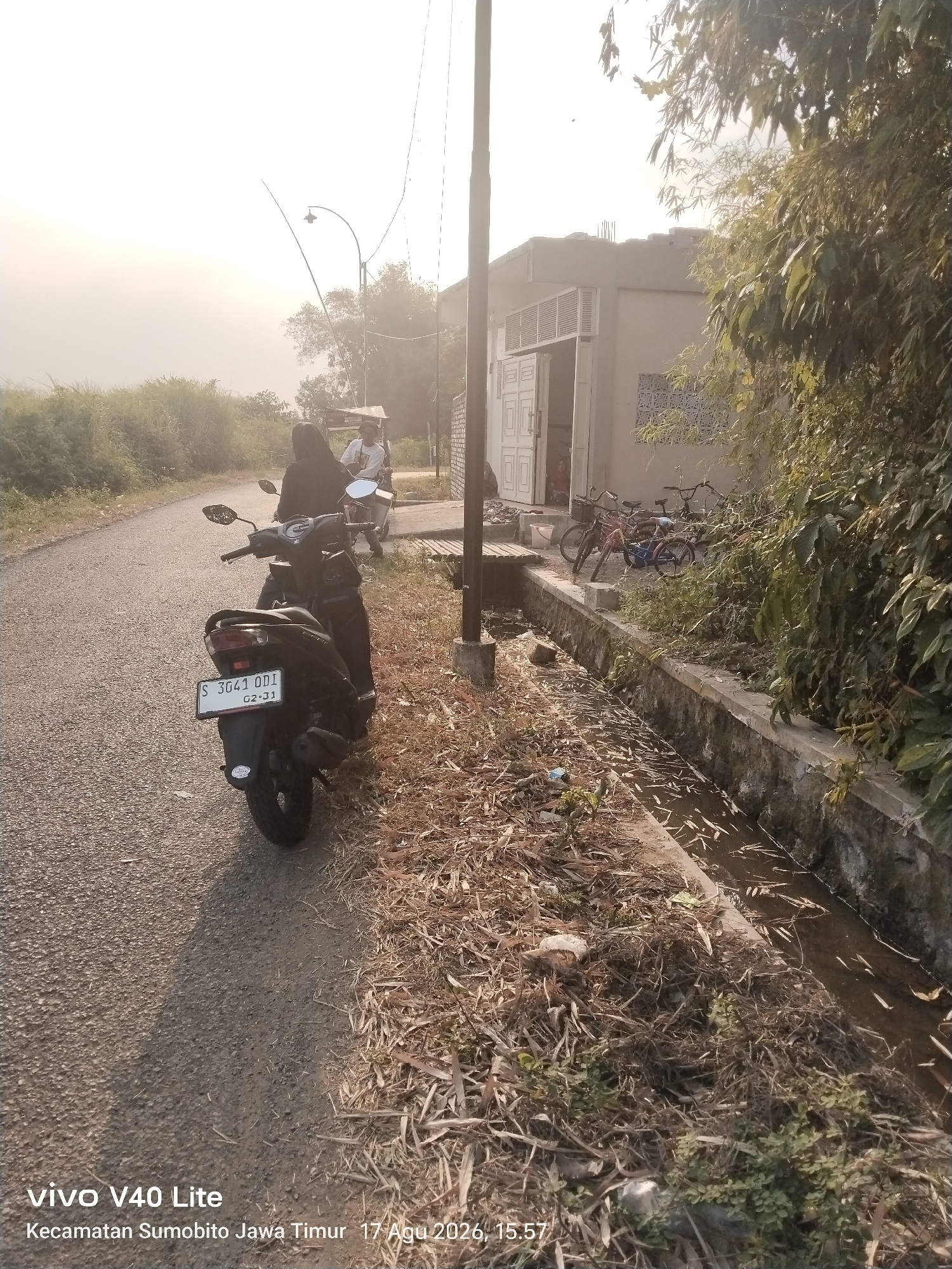 In the city of Brudu, Indonesia, on August 17, 2026, at 15:57, a scene unfolds on a paved road next to a drainage ditch. The road is lined with dry grass and foliage. On the left side of the road, a dark-colored motorcycle is parked, displaying a license plate reading "S 3041 OD1 QZ-J1". A person wearing a dark hijab is seated on the motorcycle. Further down the road, another motorcycle is visible with a rider wearing a white shirt. To the right of the road, a building with an open doorway and several windows is situated. A row of bicycles is parked near the building. A concrete structure, likely a drainage channel, runs alongside the road, filled with murky water and debris. The sky is bright and hazy, suggesting a sunny day. The image is a snapshot captured by a "vivo V40 Lite" device.