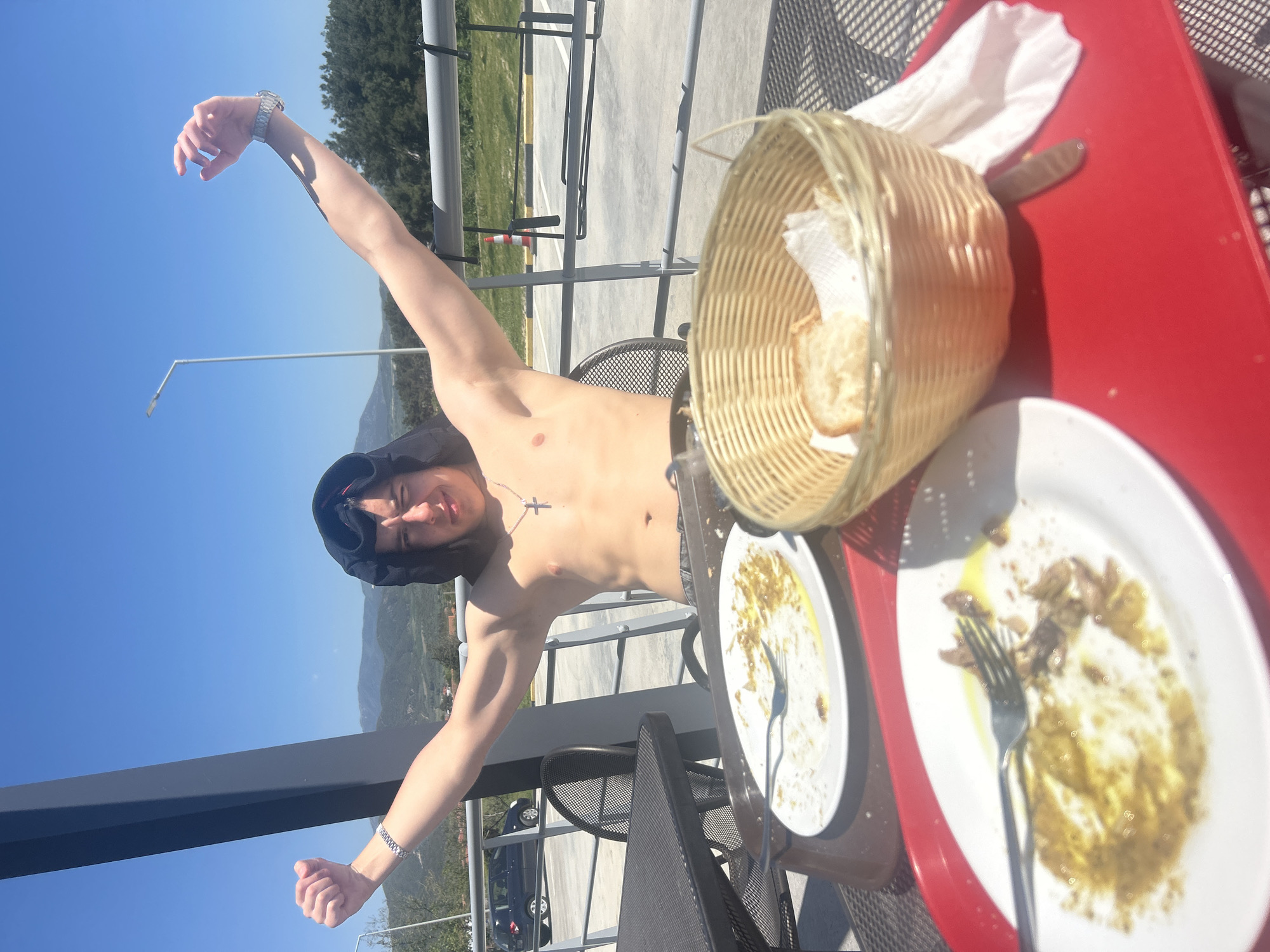 The image shows a shirtless young man wearing a dark hoodie, raising his arms in front of a railing. He is outdoors, apparently on a terrace or balcony. In the background are trees, mountains, and a clear blue sky. On a red table to the right are two plates with food remains and a fork, a bread basket with bread, and a white napkin. The setting seems to be Νέα Κερδύλια, Greece, based on the terrain.