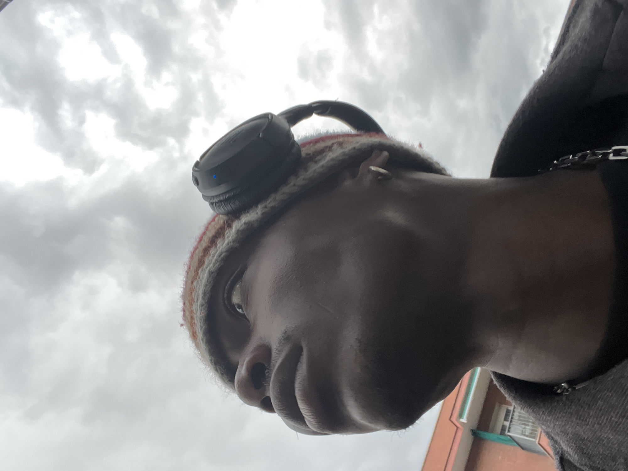 A low-angle close-up shot focuses on a person wearing a striped knit hat and black headphones. The person's face is angled towards the sky, which is filled with overcast clouds. A portion of the person's dark grey jacket and a silver chain necklace are visible on the right side of the frame. In the blurred background at the bottom right, there is a section of an orange building with white trim and a window. This image does not give enough details to conclude that it was taken in Barcelona, Spain.