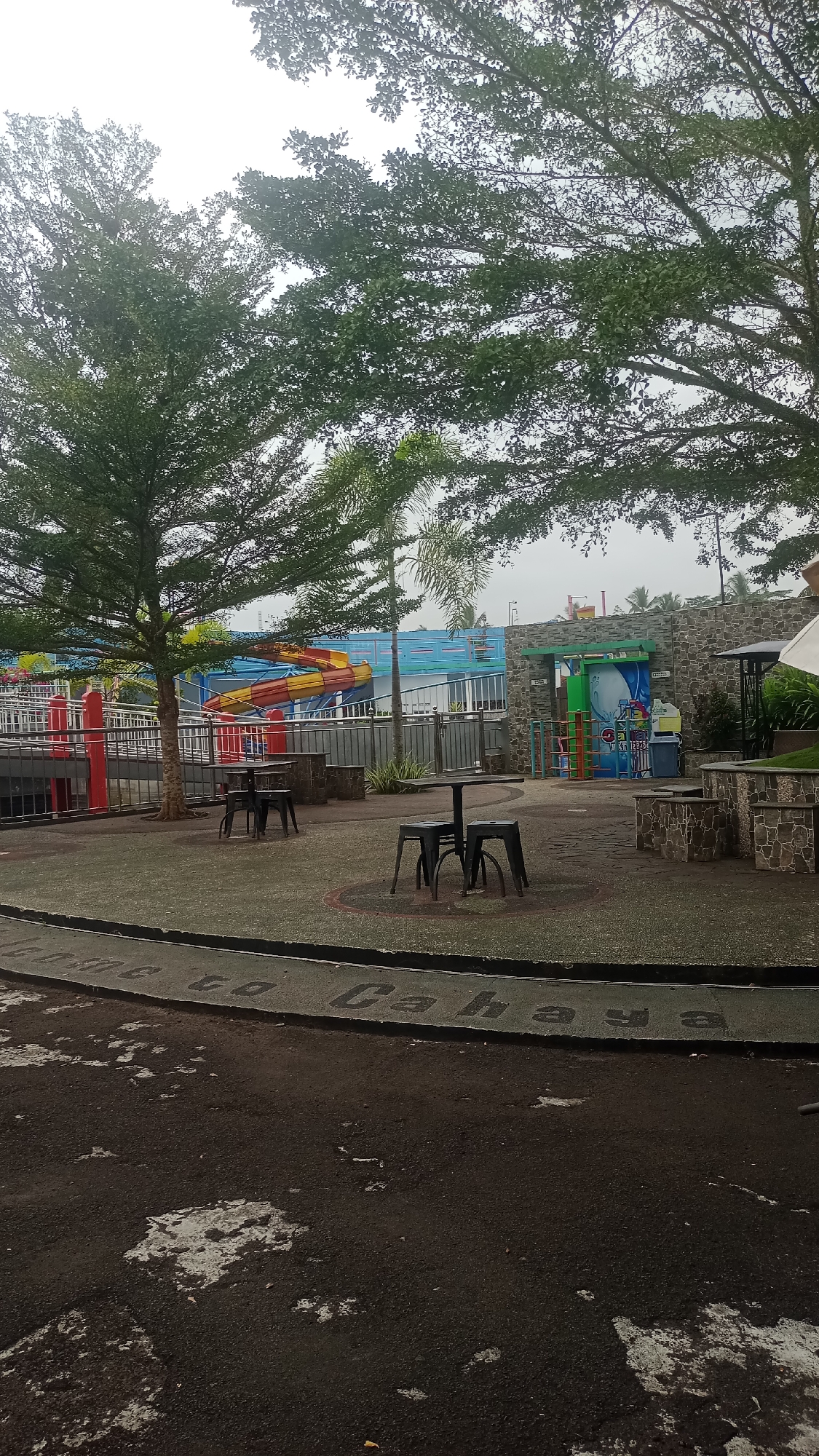 The image depicts an outdoor area that appears to be part of a water park or recreational facility, likely located in Pakemitan, Indonesia. The foreground shows a dark asphalt path with patches of lighter material, possibly concrete or worn surfaces. Curving along the path is a section of paved ground with dark, raised lettering that spells out "Welcome to Cahaya".
Beyond this path, a circular paved area contains several black plastic tables and chairs, arranged in groups of two. The ground within these circles is lighter colored, contrasting with the surrounding dark pavement. Behind the seating areas, a low stone wall encloses a grassy area.
Further in the background, a colorful water slide, primarily yellow and red, is visible. It curves downwards and appears to lead into a pool area that is partially obscured by a blue wall. Red cylindrical posts and a metal fence are also present, suggesting pathways or barriers within the facility.
To the right of the water slide, a brick structure with a green roof houses an entrance or information booth. Colorful graphics are displayed on the wall, and plastic bins are situated nearby.
Several trees with dense green foliage frame the scene. Two larger trees with spreading branches dominate the left and upper portions of the image, their leaves creating a dappled effect against the overcast sky. Smaller palm-like trees are also visible. The overall impression is of a quiet, possibly uncrowded, recreational space on a cloudy day.