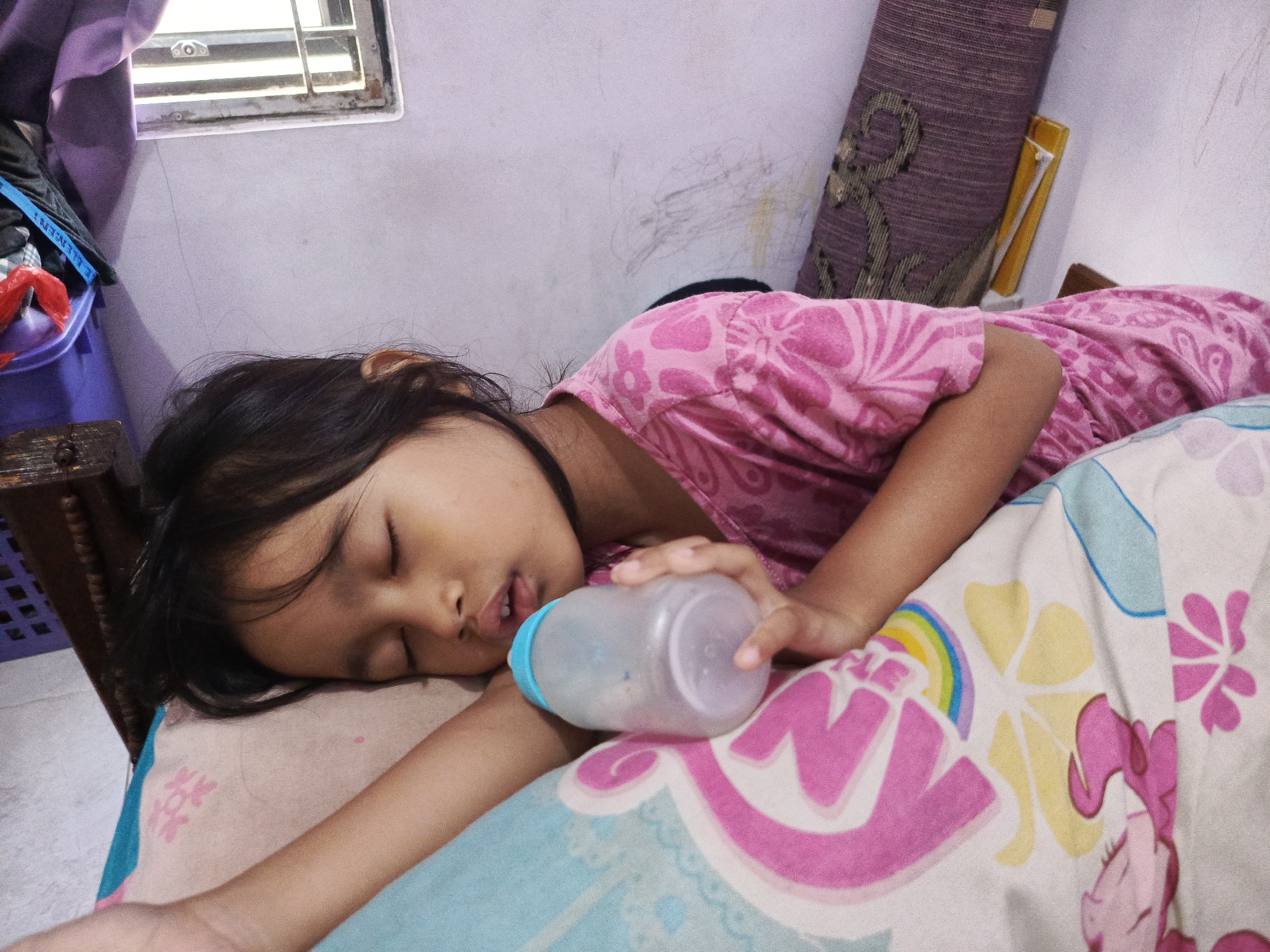 A young Indonesian girl is sleeping on a bed with a baby bottle in her hand. She is wearing a pink shirt with a floral pattern. The bed is covered with a colorful blanket featuring cartoon characters. The room appears to be a bedroom, with a window and a closet visible in the background. The overall impression is one of a peaceful, domestic scene. The location is a residential area in Ciseeng, Indonesia.