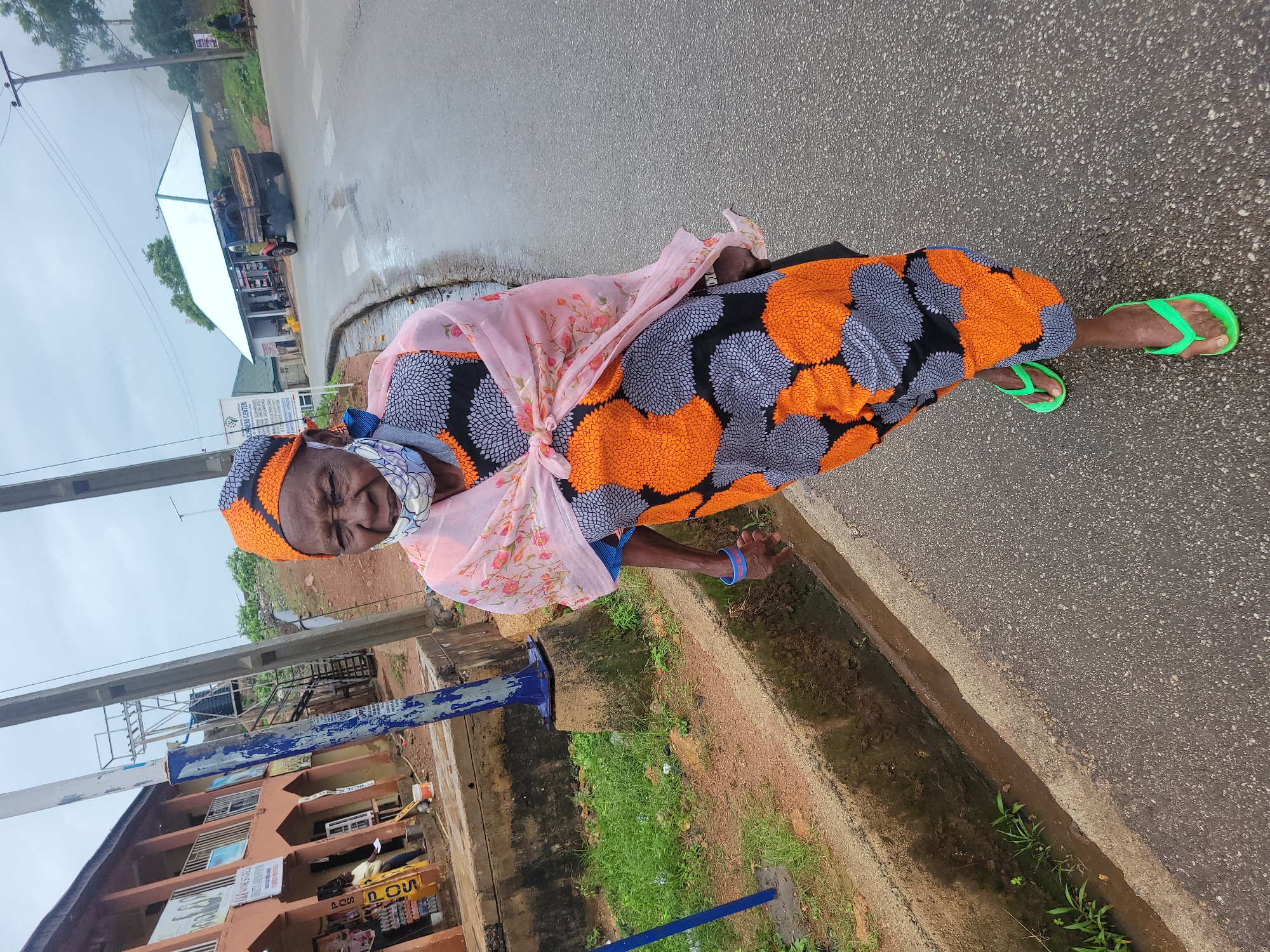 An elderly African woman wearing a brightly colored traditional outfit, a headscarf, and a face mask walks on a wet asphalt road in Jos, Nigeria. She is wearing a pink floral scarf draped over her shoulders and a dress with orange, grey, and black circular patterns. Her feet are adorned with bright green flip-flops. The woman is looking towards the camera with her eyes partially closed. To her left, there is a drainage ditch filled with water and overgrown grass. Behind her, buildings with storefronts are visible, and further in the distance, a truck is parked on the side of the road. The sky is overcast, suggesting it might be a cloudy or rainy day.