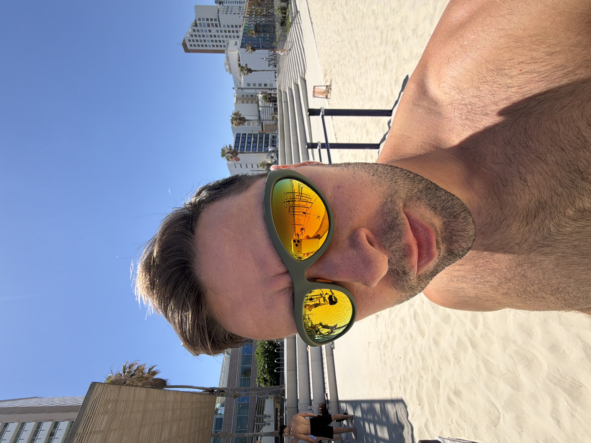 This is a vertically oriented, sunny close-up selfie of a man on a beach, likely in Tel-Aviv, Israel. The subject, a man with light stubble and shirtless, occupies the foreground, looking directly at the camera. He wears distinct olive-green framed sunglasses with highly reflective, orange-yellow mirrored lenses, obscuring his eyes.
Reflections in his sunglasses offer glimpses of the surrounding environment: the upper lens clearly shows a smartphone being held, indicating the selfie being taken, while the lower lens reflects parts of the beach infrastructure, including white architectural structures and potentially a sun umbrella or beach furniture.
The background reveals a bright, urban beach scene under a clear blue sky. A wide, sandy beach with subtle undulations stretches behind the man, leading towards several white, multi-story buildings that line the distant coastline. Palm trees are visible throughout the background. A concrete or paved walkway with steps leads down to the sand, with a few other individuals sparsely scattered on the beach, some appearing to be relaxing or walking. The bright sunlight casts distinct shadows, suggesting a clear day during mid-morning or afternoon. No visible text is present in the image.