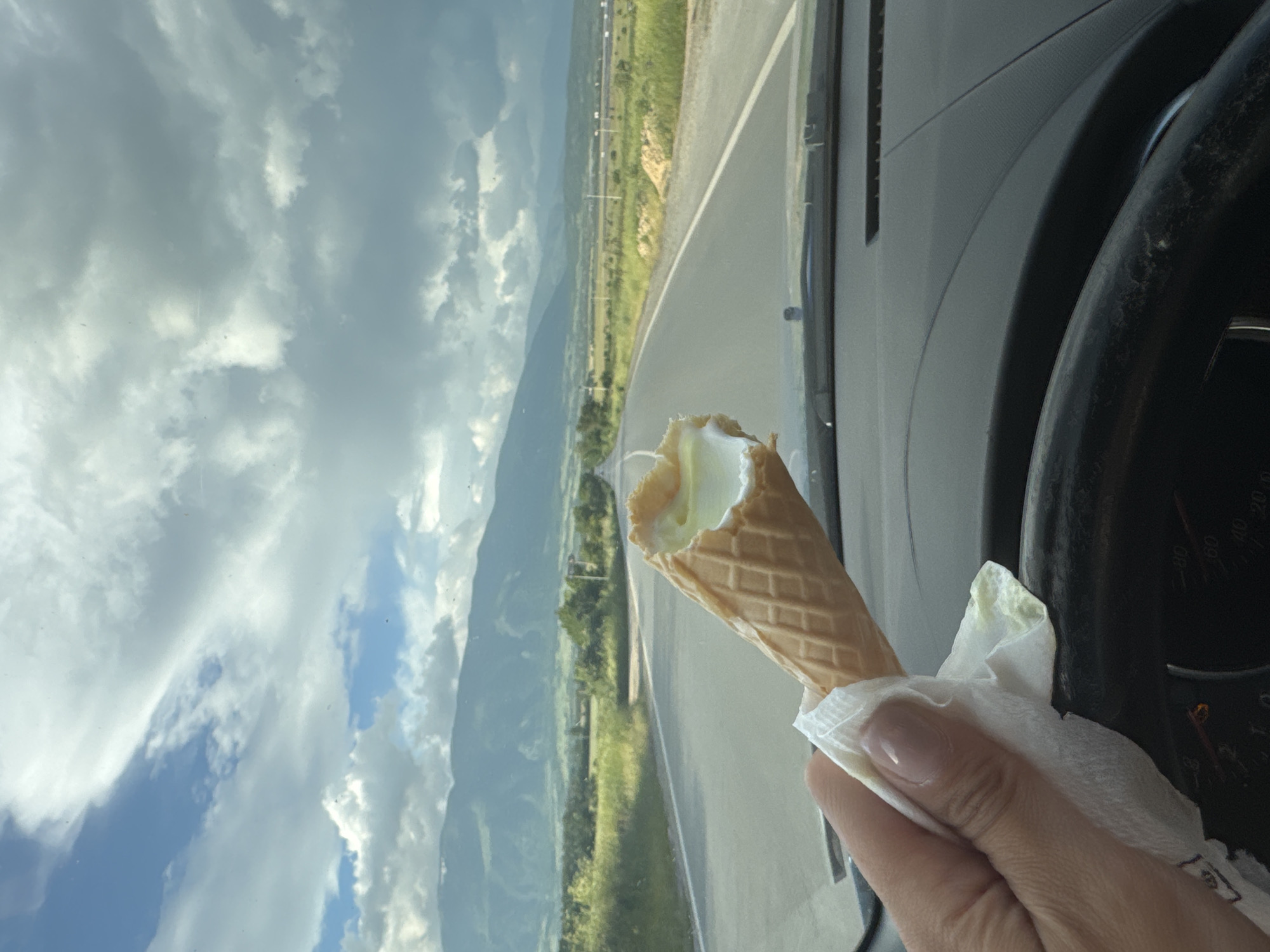 An ice cream cone with a bite taken out is being held in a hand inside a car. The background shows a road and landscape with mountains, visible through the car's windshield. The car's steering wheel is also visible. The location appears to be in or near Vedrare, Bulgaria, based on landscape characteristics.