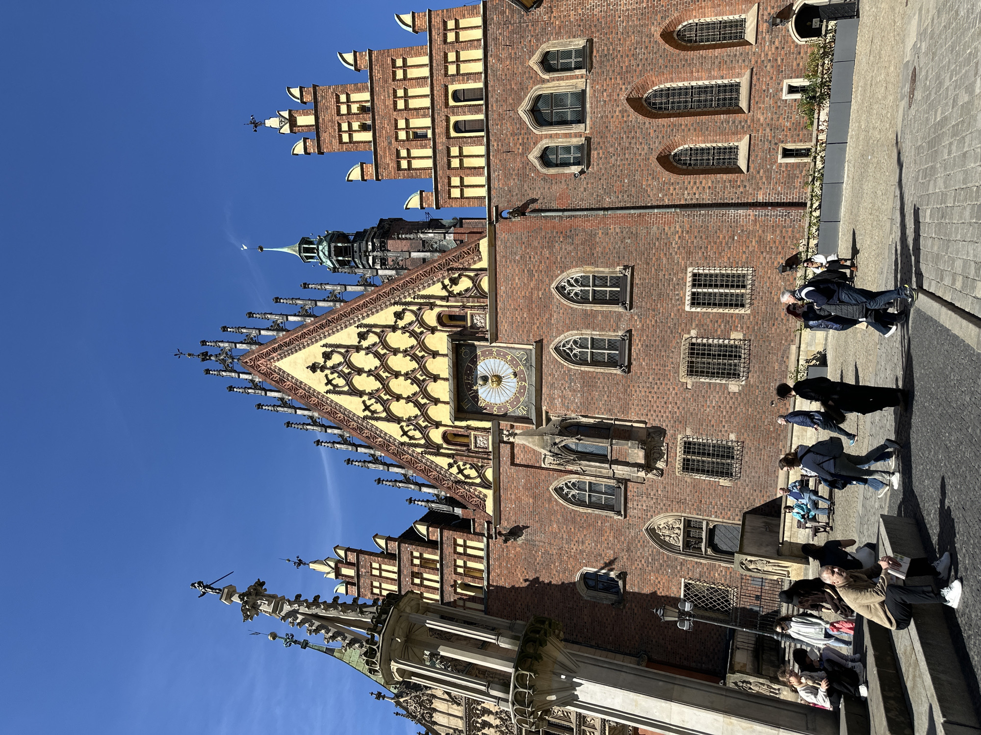 The image depicts the Old Town Hall in Wrocław, Poland, taken on a sunny day. The building's architecture features brickwork, pointed windows, ornate gables, and a clock tower. Several people are present on the sidewalk in front of the building, including a group sitting on a ledge and others walking. The sky is clear and blue.