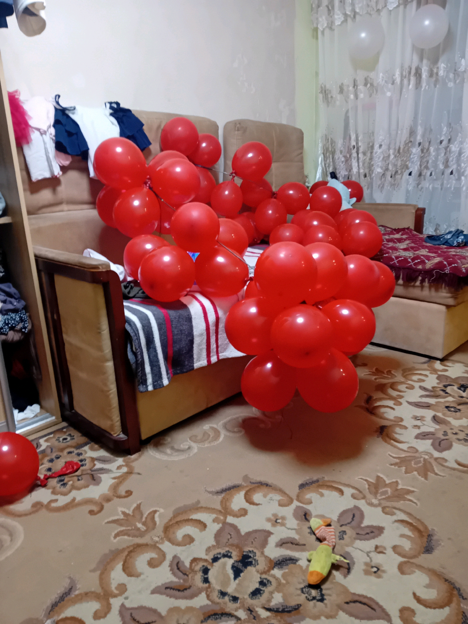 The image depicts a domestic scene within an apartment in Chuhuiv, Ukraine, characterized by a festive atmosphere indicated by numerous red balloons. The primary subjects are these balloons, clustered and scattered throughout the room. A sofa occupies a central position, draped with a striped blanket and a pile of clothes. To the left, a closet or wardrobe is partially visible, containing more clothing. The floor is covered by a patterned carpet. In the background, a curtained window is visible, with two white balloons floating near it. A small, stuffed toy lies on the carpet. The lighting suggests an indoor environment, possibly evening. No people are present in the frame.