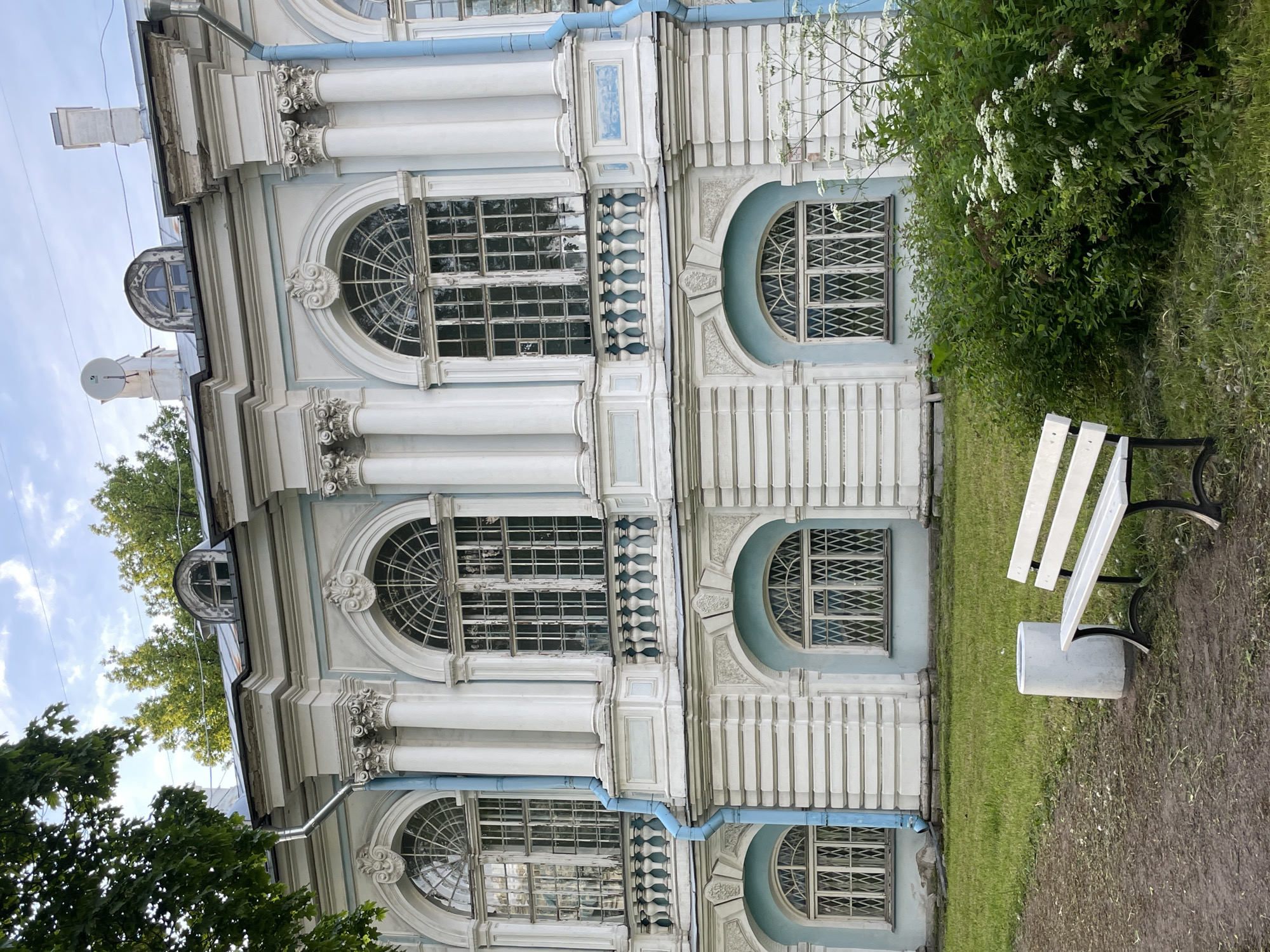 This image captures the facade of a large, two-story building in Unknown, Russia, under a partly cloudy sky. The building exhibits a neoclassical architectural style with a light blue and white color palette. The upper story features three prominent arched windows with multi-pane white grilles, flanked by white columns with ornate capitals. Decorative cartouches with shell motifs are positioned above the central upper windows. White balustrades are present below the two rightmost upper windows. The lower story contains arched window recesses, also painted light blue, with windows secured by diamond-patterned white grilles. Fluted pilasters separate the lower-story window bays. Two light blue downspouts run vertically down the facade. The roofline includes multiple dormer windows, some arched and one circular, along with several chimneys and a visible satellite dish. The building shows signs of aging, including chipped paint, weathered window frames, and some damaged glass panes.
In the foreground, a dirt path occupies the left side, transitioning into a grassy area. A white park bench with black metal legs is positioned to the right of a cylindrical grey concrete trash bin. Green foliage, including large deciduous trees in the background and a dense cluster of shrubs with small white flowers in the midground, frames the lower right portion of the building. No individuals are present in the scene.