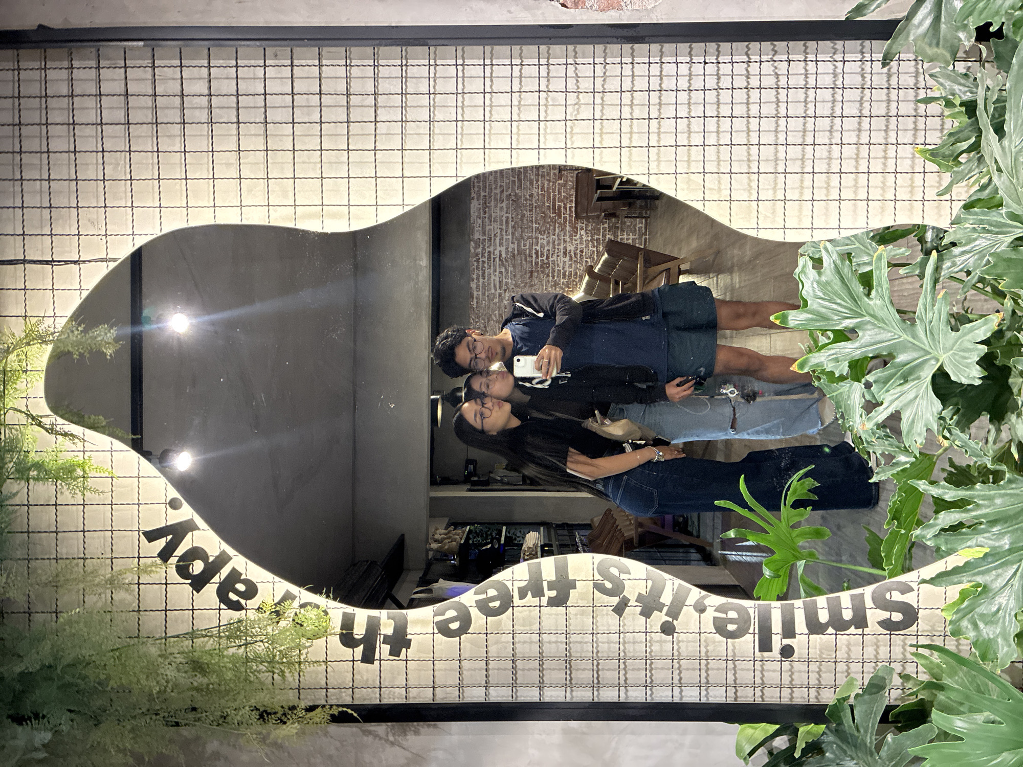 The image depicts a couple taking a selfie in front of an ornate mirror. The mirror has a wavy, irregular shape and is framed by a wire grid. The words "smile, it's free therapy" are written along the mirror's edge. The reflection shows the couple standing close together, holding a phone up to take the picture. The surroundings in the reflection include brick walls, tables, and chairs. Outside the mirror's reflection, there is greenery, including potted plants with large leaves, adding to the decorative setting. The location is likely a cafe or restaurant in San Andrés Cholula, Mexico.