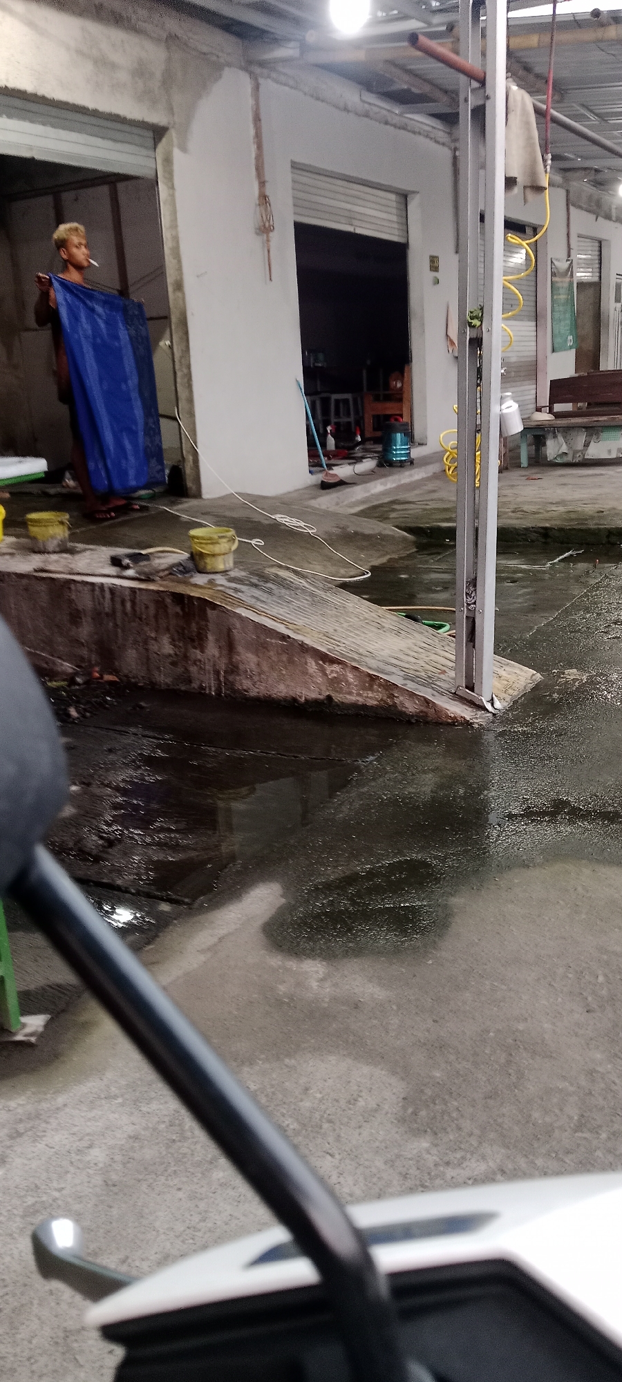 A person with blonde hair is standing in an open doorway, holding a blue tarp and smoking a cigarette. They are in what appears to be an outdoor car wash or workshop area in Maguwoharjo, Indonesia. The ground is wet and appears to be flooded in some areas. There are several yellow buckets on a concrete ramp leading into the doorway. A metallic structure with yellow coiled hoses is visible to the right, along with a wooden bench. The scene is lit by overhead lights, casting reflections on the wet surfaces.