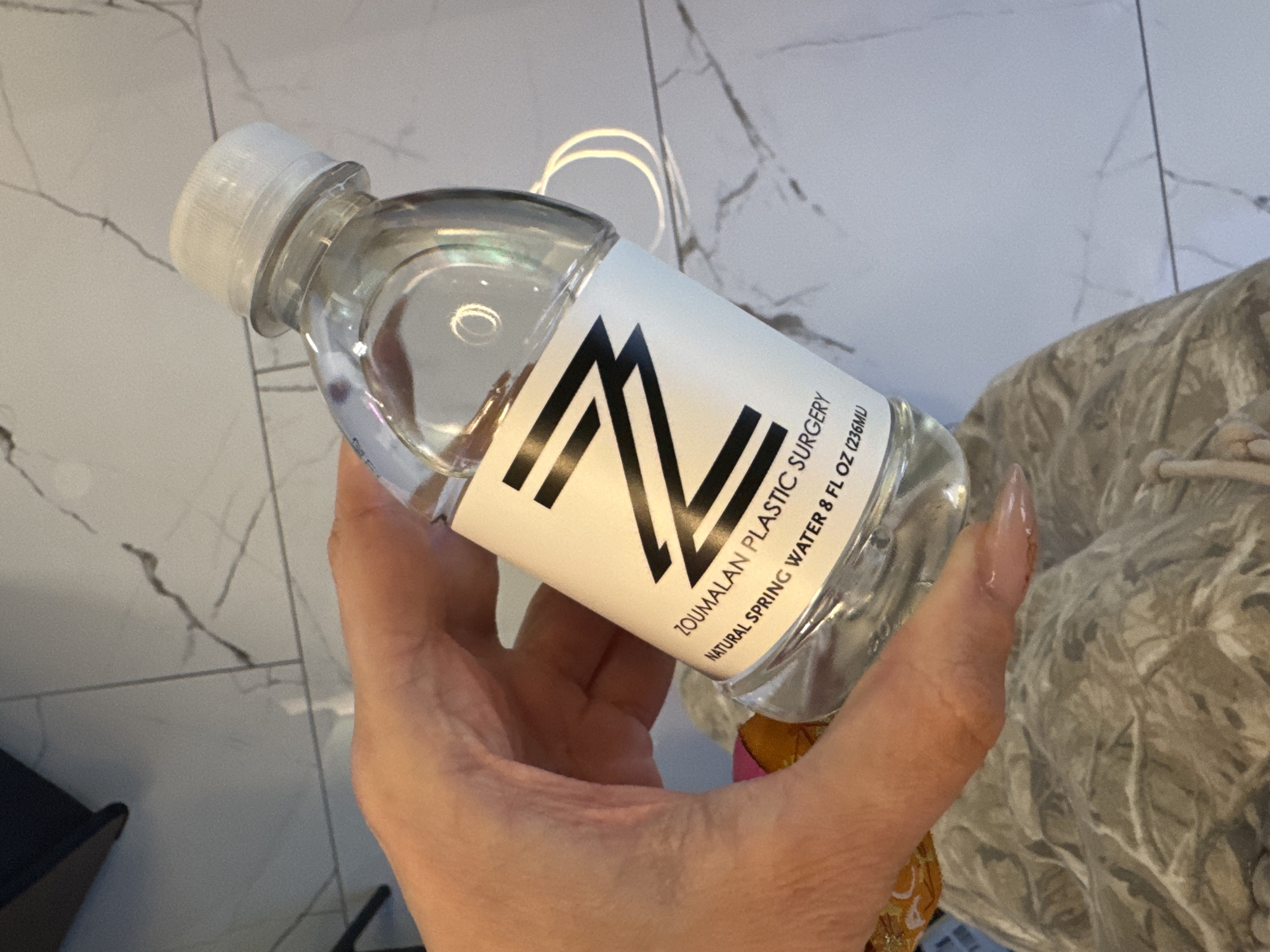 A hand holds a small clear plastic water bottle with a white cap and a white label featuring a large black geometric logo of two intersecting "V" shapes and the text "ZOUMALAN PLASTIC SURGERY NATURAL SPRING WATER 8 FL OZ (236ML)". The background includes a marble-patterned tile floor and a partially visible beige patterned soft material like fabric or upholstery. The location is identifiable by the brand of water which identifies a business in Beverly Hills, United States of America.