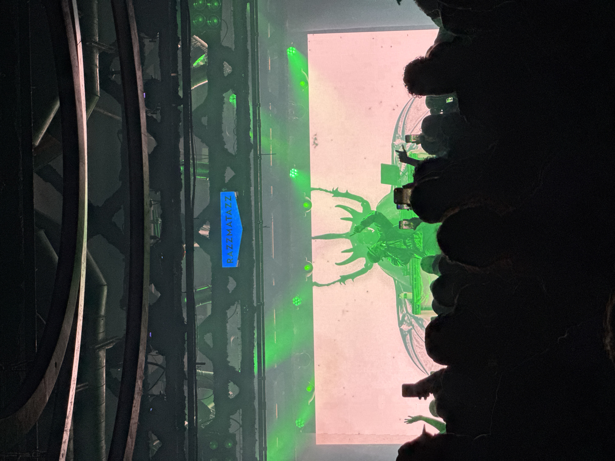 The image depicts a live concert or performance venue. On the left is structural support with metal beams and pipes. To the right is a large rectangular screen projecting an image of a green, fantastical insect-like creature with visible detail. Green spotlights illuminate the stage and the projection. The audience, partially visible in the dark foreground, is holding up mobile phones, seemingly recording or photographing the performance. A sign with "RAZZMATAZZ" printed vertically is attached to the structural support on the left. This suggests the location is Razzmatazz, a well-known concert venue in Barcelona, Spain.