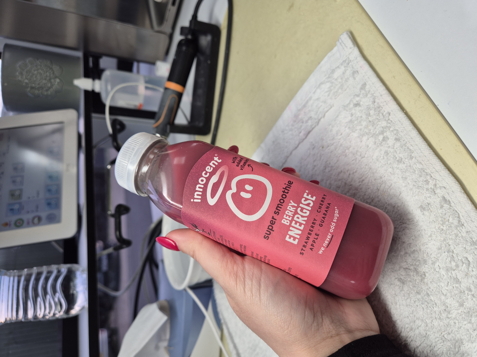 A hand holds a bottle of Innocent super smoothie "Berry Energise" drink. The drink contains strawberry, cherry, apple and guarana. The label is pink and has the Innocent logo of an apple with a halo. The background is blurred, but appears to include a white towel, a tablet device, and some other equipment. The location is unknown.