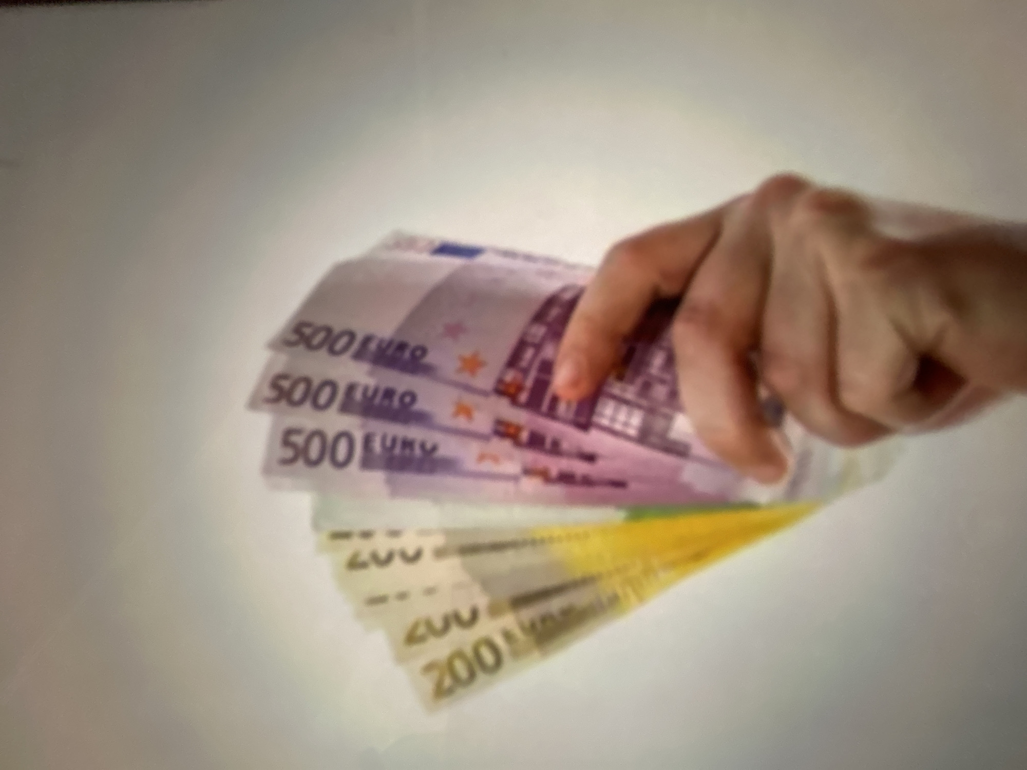 A hand is shown holding several euro banknotes. The banknotes are fanned out, with the denominations of 500 and 200 euros visible. The 500 euro notes are purple, and the 200 euro notes are yellow and green. The background is a plain, light-colored surface, suggesting an indoor setting. There are no discernible location cues or indications of time of day or weather. The image does not convey any specific emotion. The text "500 EURO" and "200 EURO" is clearly visible on the respective banknotes. The overall impression is a close-up shot focusing on the presentation of money.