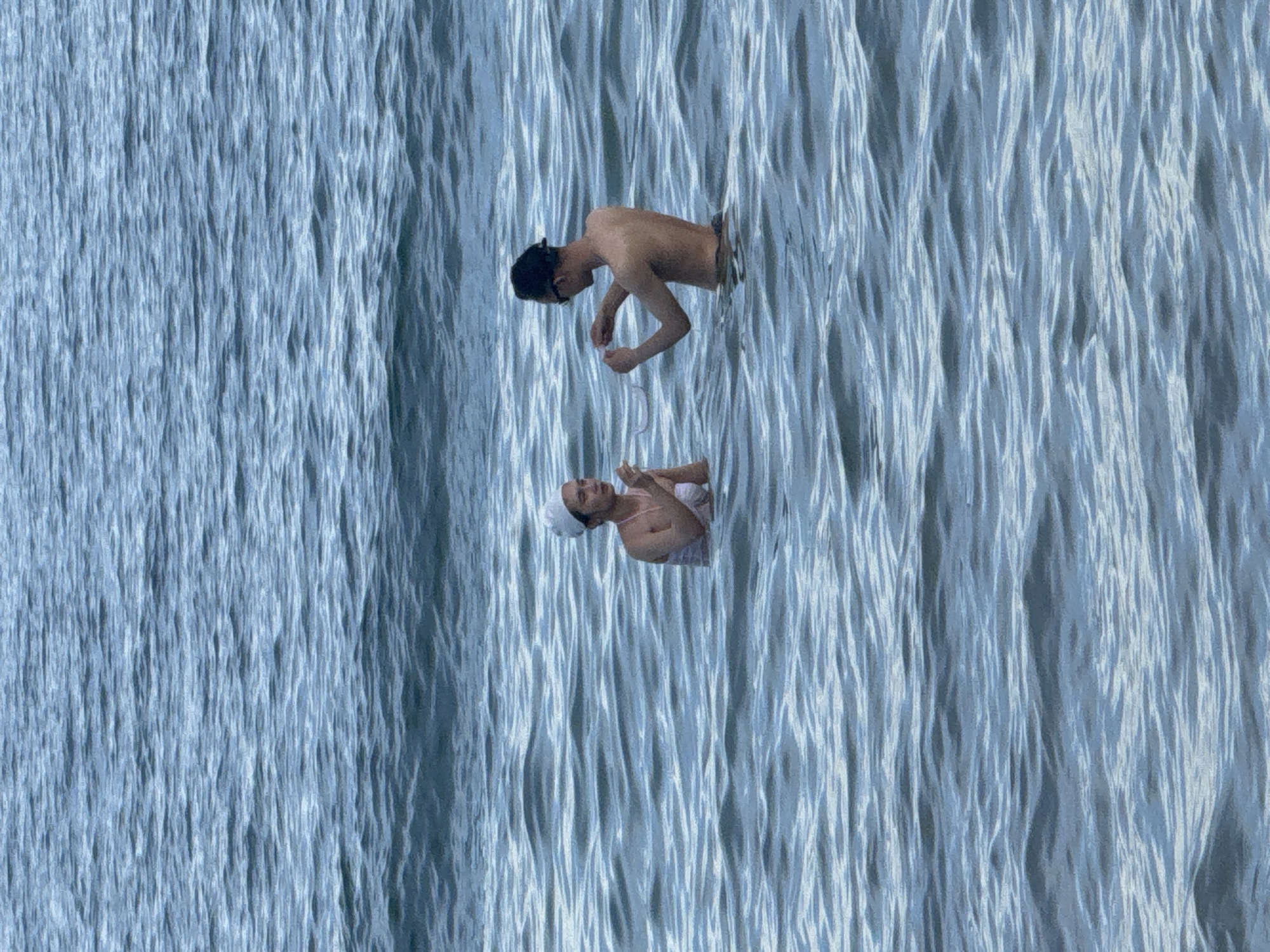 Two people are standing in the water. The person on the left is a woman wearing a white headscarf and a light-colored swimsuit. She is holding something in her hands, bringing it to her mouth. The person on the right is a man with dark hair, wearing sunglasses. He is also holding something in his hands and appears to be interacting with the woman. They are both partially submerged in the water, which is calm with gentle ripples. The background is the open water, with no land or structures visible. The setting appears to be a body of water, likely the sea or a large lake, in Phu Thuy Ward, Vietnam.