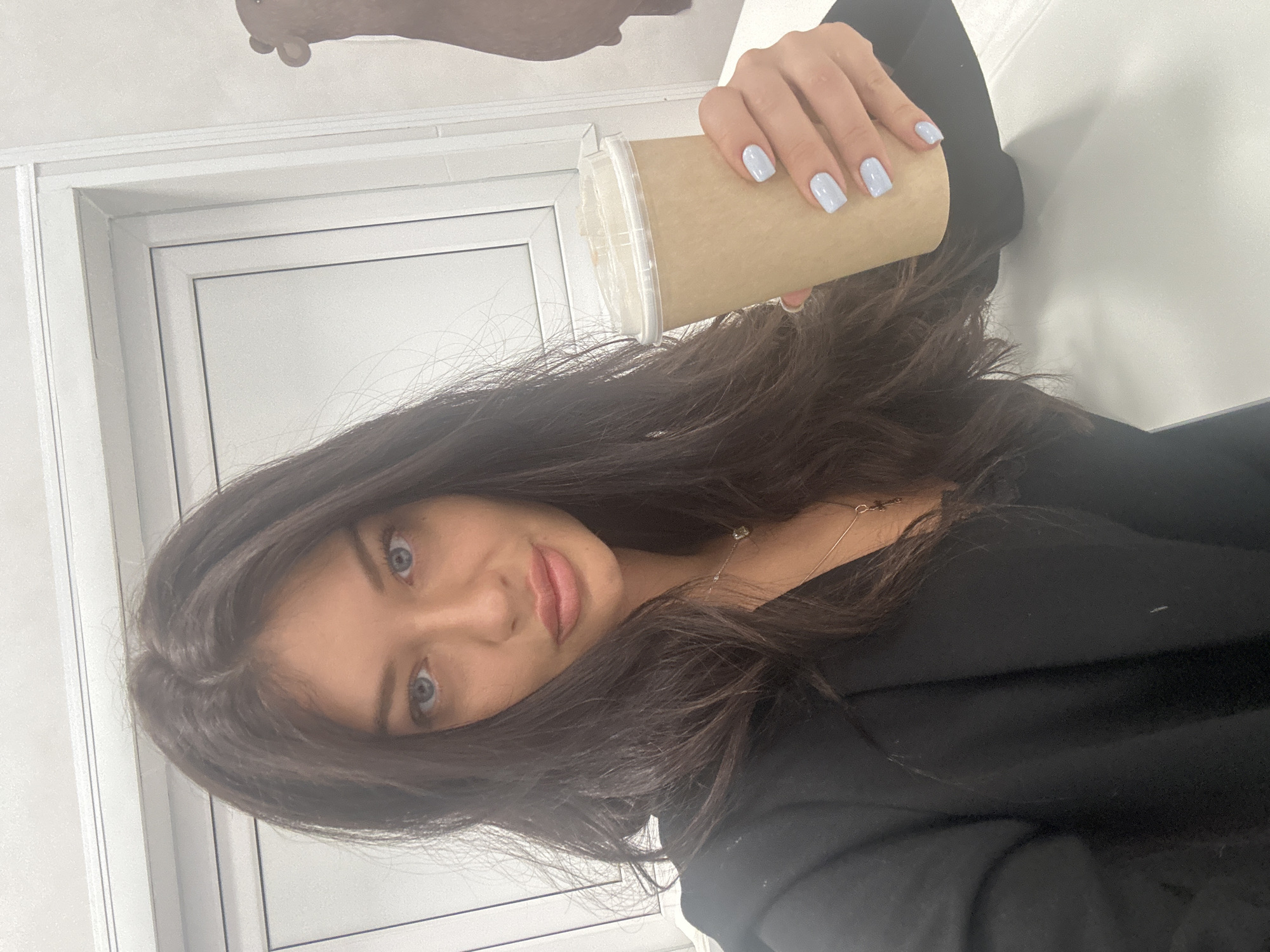 An image, rotated 90 degrees counter-clockwise, features a young woman looking directly at the camera with a slight smile. She has long, dark brown hair that falls over her shoulders and partly obscures her face, revealing her blue eyes. She is wearing a dark, likely black, top or jacket, and a delicate silver necklace with a small pendant is visible around her neck.
In her right hand, she holds a light brown, disposable paper cup with a clear plastic lid, suggesting it contains a beverage like coffee or tea. Her fingernails are painted with a light blue or periwinkle polish. The background consists of a plain white wall and a white door or window frame, indicating an indoor setting. The lighting is bright and even. No discernible text is visible on any objects or in the background.