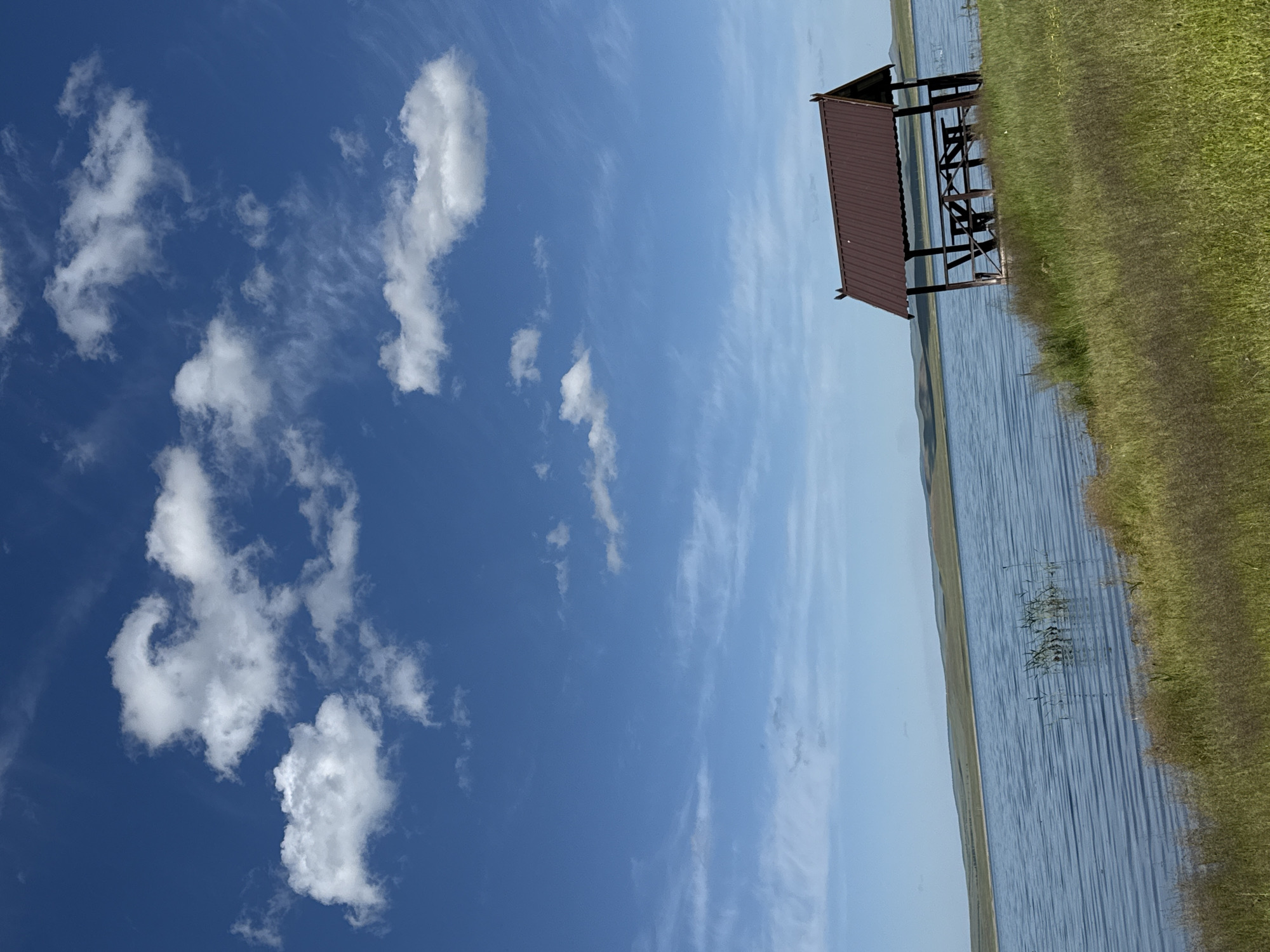 The image displays a serene lakeside scene with a boathouse. The sky is a vibrant blue, dotted with scattered white clouds, suggesting a fair-weather day. The sun appears to be high in the sky, casting a bright light on the scene, indicating it is likely daytime.
The foreground is dominated by lush green grass that slopes down towards the water. The lake itself is calm, with gentle ripples on its surface reflecting the blue of the sky. In the middle ground, a rustic boathouse stands on the water's edge. It has a dark, possibly brown or maroon, metal roof supported by wooden beams, and an open-sided structure beneath. The boathouse appears to be empty, with no boats or people visible.
In the background, the land is visible as a gently rolling hillside, some of which is covered in dry grass, and a distant horizon line where the land meets the sky. The overall mood of the image is peaceful and tranquil, evoking a sense of quiet solitude in nature. There is no visible text in the image.