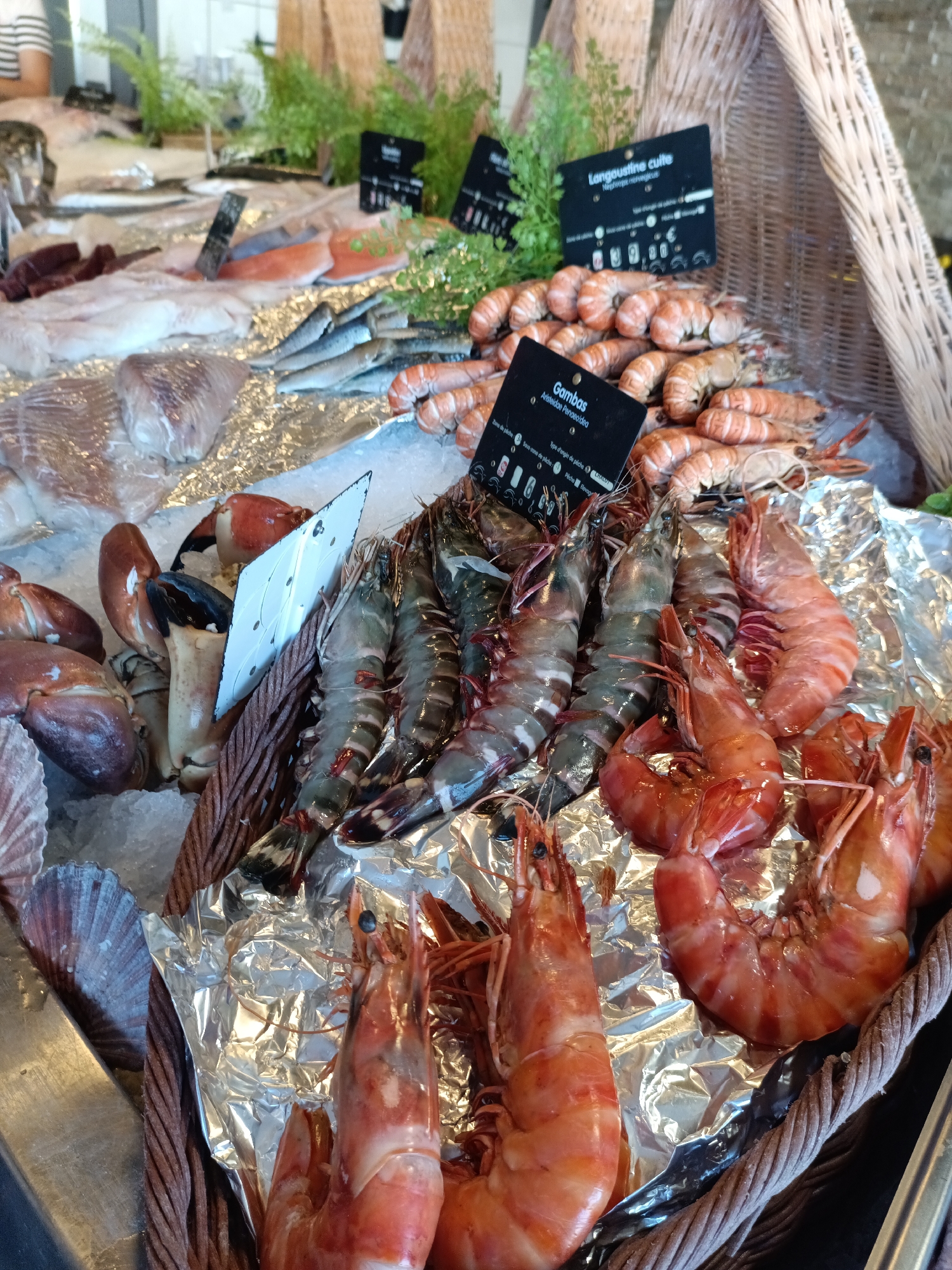 The image shows a display of fresh seafood at a market in Le Neubourg, France. The seafood is arranged on ice and in baskets, with several types of fish and crustaceans visible. Prominently featured are large prawns (gambas) and langoustines, some of which are cooked and have a reddish-pink hue. Other seafood items include what appear to be crab claws and various types of fish fillets. Small black signs with white text are placed among the seafood, likely providing information such as the type of seafood and its price. The overall setting suggests a bustling market environment with an emphasis on fresh, high-quality seafood. The background shows woven baskets and some greenery, further contributing to the market atmosphere.