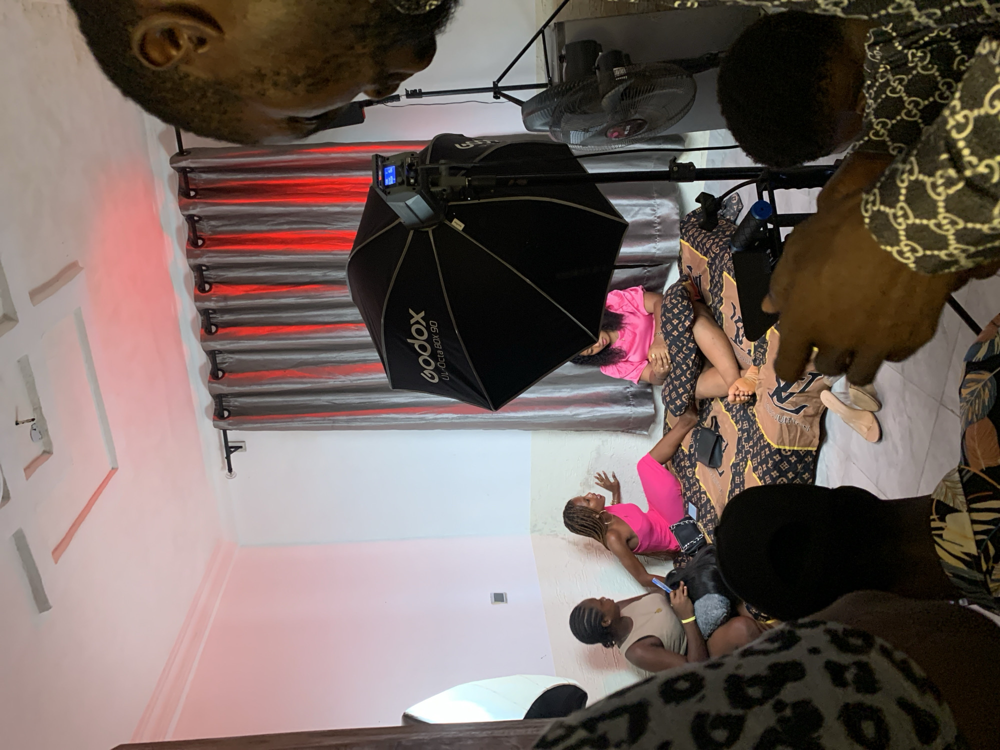 The image, taken indoors in what appears to be a studio or a styled room in Ikorodu, Nigeria, captures a photoshoot or video recording session. Several individuals are present, with the main subjects being two women on a bed or lounge area. One woman, dressed in a bright pink top, is sitting up on the bed, while another in a matching pink jumpsuit is lying on her side, posing. The bed is covered with patterned bedding resembling the Louis Vuitton monogram.
In the foreground, partial views of other individuals are visible. One person's head is seen in the top left, while another, wearing a black and white patterned shirt (reminiscent of Gucci), is seen from the back on the right side. Further down, another person is seated on the floor, looking at a mobile phone.
The setup includes professional lighting equipment: a large black Godox-brand octagonal softbox, partially revealing text like "Godox UL-60" and "UL150 R 0-100%," is positioned over the subjects. Two black standing fans are visible in the background, suggesting a warm environment. The room features white walls, with some decorative rectangular indentations, and gray or silver curtains illuminated from behind by red ambient lighting, casting a reddish glow on the surroundings. A black handbag rests near the models, and footwear is seen on the floor beside the bed. The overall scene suggests a creative production activity taking place, likely in the evening or night due to the controlled artificial lighting.