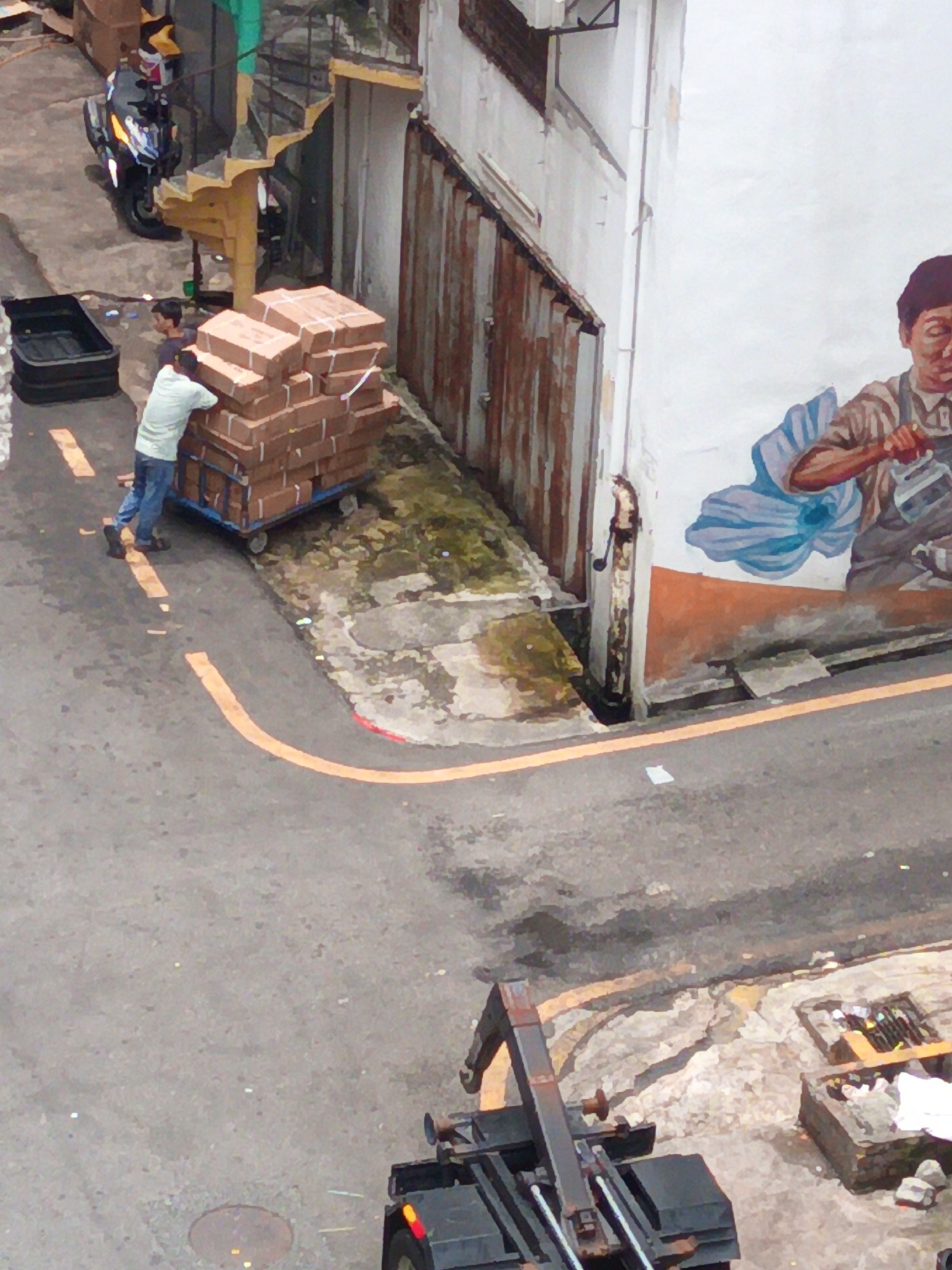 An overhead, slightly angled view shows an urban scene in Kuala Lumpur, Malaysia. In the foreground, a person in a light blue shirt and dark pants is pushing a cart laden with stacks of cardboard boxes. The cart has blue metal supports. To the left of the cart, a dark plastic bin sits on the asphalt. Behind the cart, a motorcycle is parked, facing away from the viewer, near a spiral staircase that curves upwards.
A mural adorns the white wall of a building in the background. The mural depicts a person, possibly a laborer, with brown hair and wearing a striped shirt and overalls, holding a tool. A large blue floral element is also part of the mural. The building has a corrugated metal siding on its lower section. The ground is asphalt, with faded yellow markings indicating a pathway or lane. There is some patchy green moss or vegetation growing near the wall and sidewalk. In the far right background, a small section of what appears to be a construction or scaffolding element is visible, along with a brick structure containing debris. The overall impression is one of a busy, working-class urban environment.