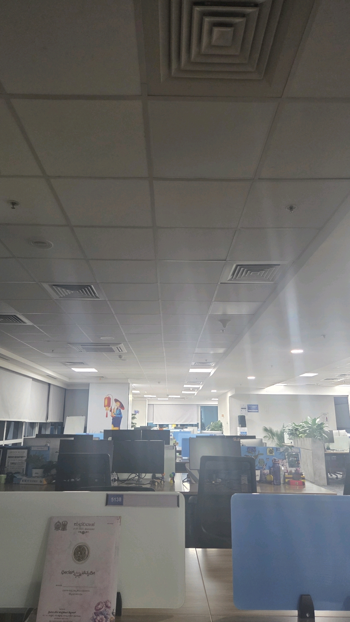 The image depicts an indoor scene, likely an office space, with a focus on the ceiling and a row of cubicles. The ceiling is constructed of modular tiles and features several ventilation grilles and light fixtures. In the foreground, a stack of paper, possibly documents or a pamphlet, rests on a desk. The document has text in a script that appears to be Kannada, indicating a location in India, likely Bengaluru, given the prevalence of the script there. Behind the document, multiple computer monitors are visible, suggesting a work environment. Further in the background, more cubicles and office furniture can be seen, along with a wall graphic featuring a stylized illustration of a person holding a lantern. The overall impression is of a functional, modern office.