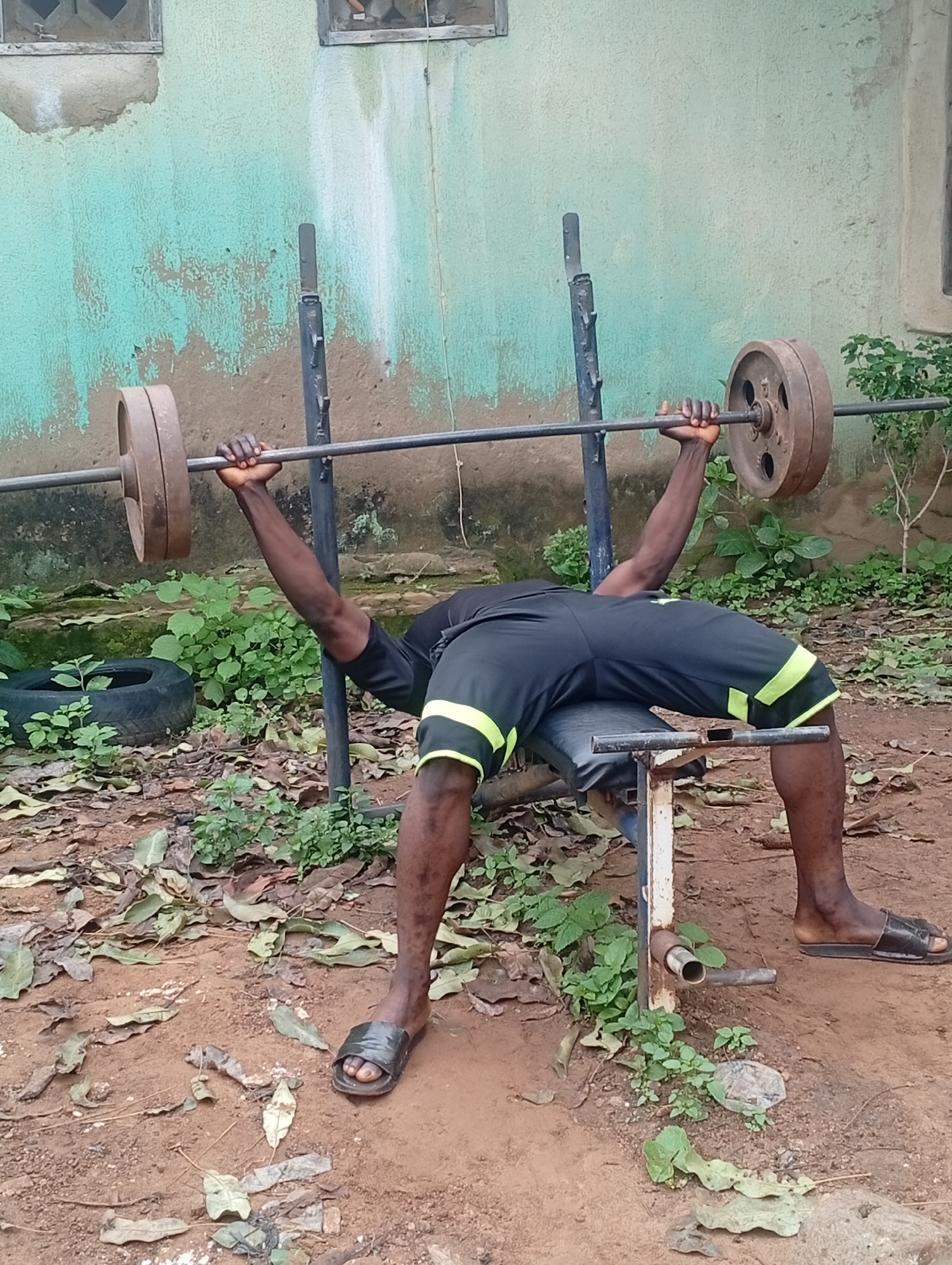 An individual is performing a bench press exercise outdoors in Akwanga, Nigeria. The scene is set against a weathered, light teal-colored wall with visible patches of exposed concrete and some green vegetation growing near its base. In the foreground, the individual is lying on a rusty white weight bench. They are wearing black shorts with neon yellow stripes along the sides and black flip-flops. The individual is lifting a barbell loaded with two heavy, circular weight plates, one on each end of the bar. The barbell rests on the individual's chest, and their arms are bent at the elbows, indicating the midpoint of the bench press motion. The ground around the bench is covered with reddish-brown dirt and scattered dry leaves, with more green plants visible in the background. A used tire is partially visible to the left of the bench. The lighting suggests it is daytime, possibly overcast or shaded.