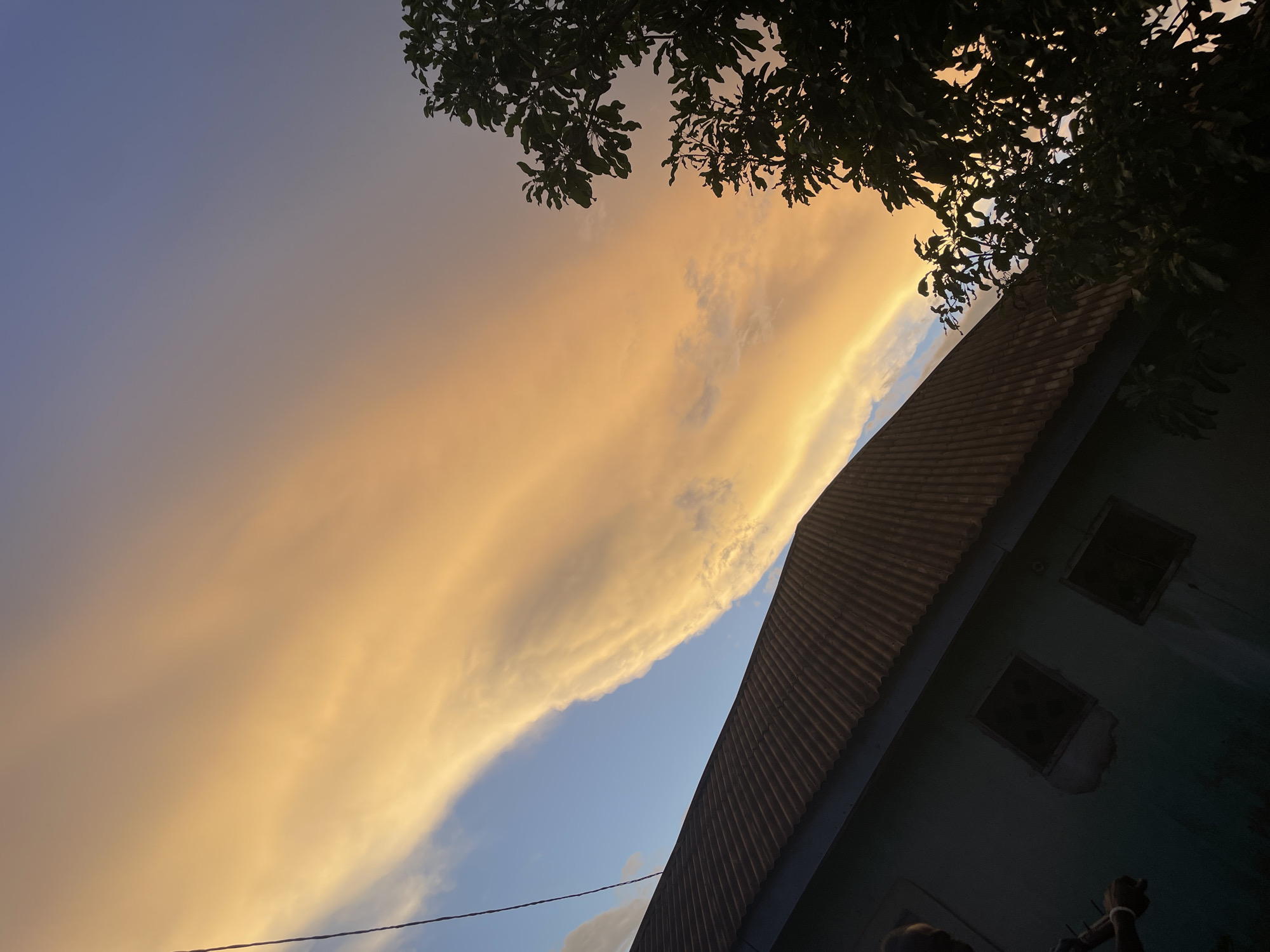 The image depicts an outdoor scene at twilight in Akwanga, Nigeria. The sky transitions from a deep blue and purple in the upper portion to a bright orange and yellow hue closer to the horizon, indicative of a low sun angle. A prominent, elongated cloud formation, illuminated with intense golden-orange light, stretches diagonally across the sky. In the lower-left quadrant, a section of a building is visible, featuring a dark, corrugated metal roof and walls of a muted teal color, largely obscured by shadow. Two distinct, unlit rectangular window openings are visible on the building's wall. A dark utility cable extends diagonally from the upper-left corner of the frame, passing in front of the clouds and descending towards the left side of the building. Silhouetted trees with dense, dark foliage occupy the right side of the image, obscuring a portion of the sky. A person's upper torso and arms are partially visible in silhouette on the far-left, with an object discernible on the left wrist.