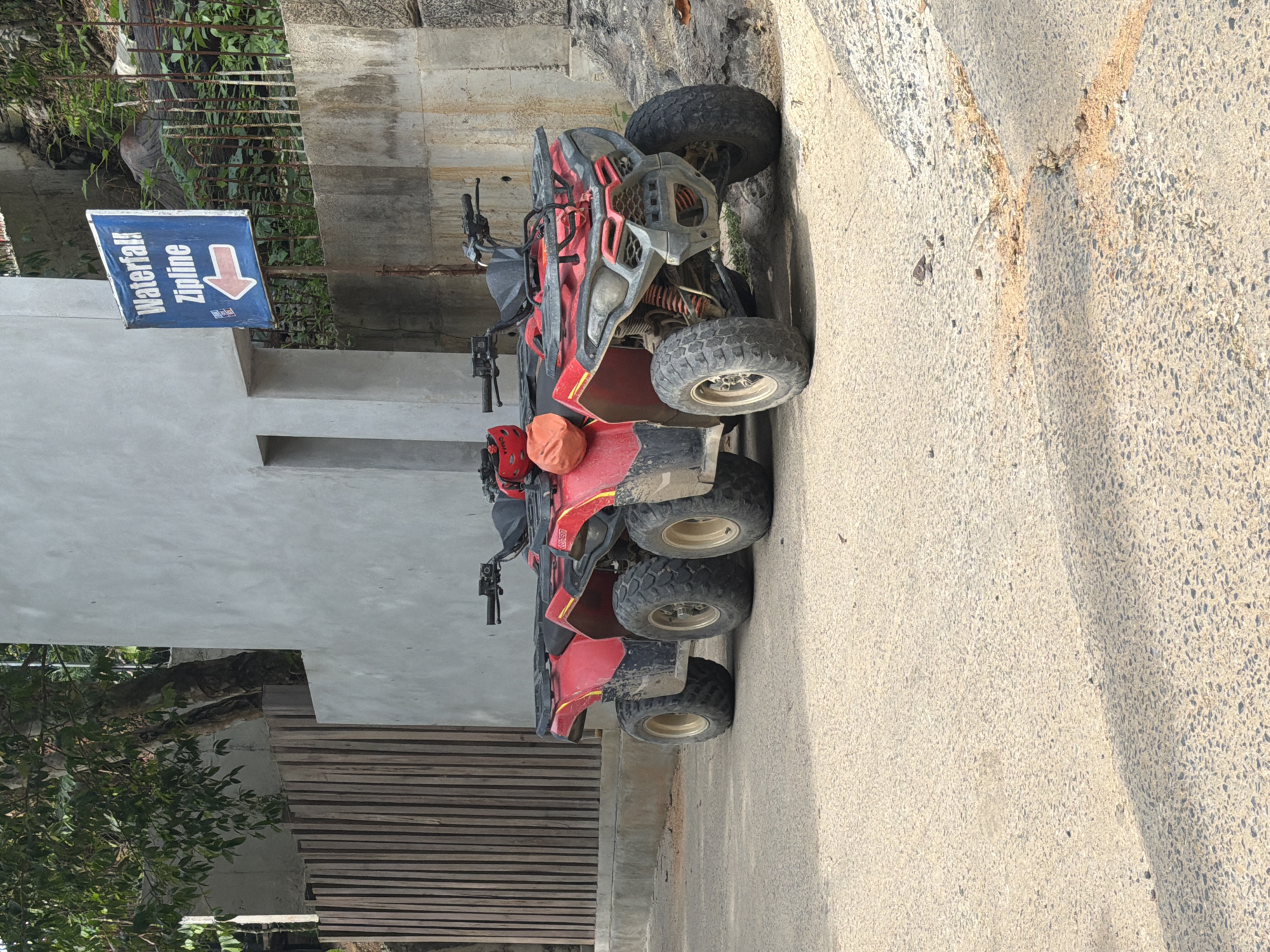 A six-wheeled, red ATV is parked on a paved road in front of a building and a wooden fence. The ATV has a black seat, black handlebars, and large tires with gold rims. A red helmet and an orange bag are attached to the back of the ATV. A blue sign with white text and a red arrow is mounted on a pole to the right of the ATV. The sign reads "Waterfall Zipline" and the arrow points to the left. The environment appears to be tropical, with lush green foliage visible in the background. The building is modern and has a light-colored, textured exterior. The ground is paved and shows some signs of wear and tear, with loose dirt and pebbles scattered across the surface. The overall scene suggests a location that offers adventure activities. This scene is located in Koh Samui, Thailand.