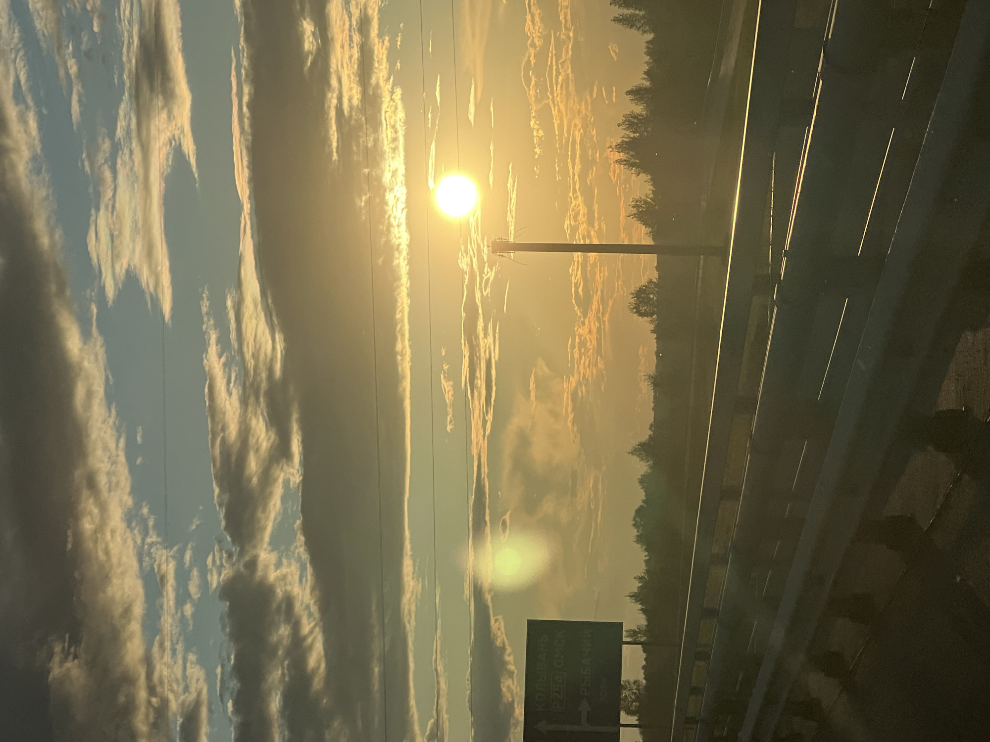The image depicts a sunset scene from a moving vehicle, likely on a highway or a road with a guardrail in the foreground. The sky is filled with dramatic clouds illuminated by the setting sun, which is a bright, glowing orb towards the right side of the frame. The light from the sun casts a warm, golden hue across the sky and the landscape. In the lower left of the image, a green road sign is visible, indicating directions to "KOLYVAN", "P254 OMSK", and "RYBACHIM". The landscape beyond the road appears to be a forest or a line of trees silhouetted against the vibrant sky. There are no people or explicit actions visible in the image, creating a sense of travel and observation of the natural environment. The location is identified as the city of Mochishche, Russia, suggesting the scene is captured on a road leading to or from this area.