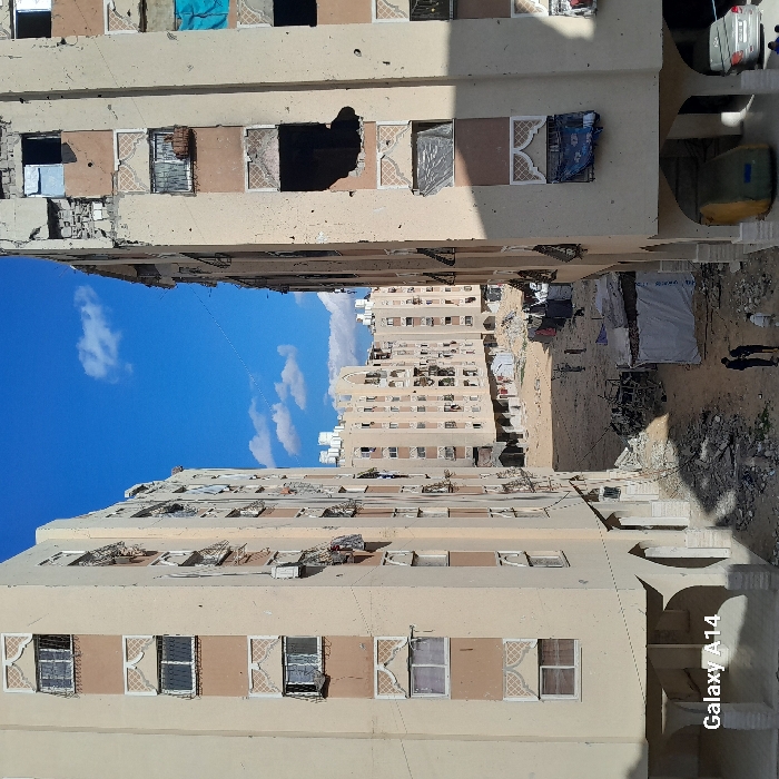 This image depicts a scene in the city of Khan Yunis, Palestinian Territory, characterized by extensive structural damage under a clear blue sky with scattered white clouds. The view looks down an open area framed by two multi-story, light-colored residential buildings, both exhibiting significant damage.
The building on the left shows numerous small impact marks and partially opened or damaged windows, some with clothes or fabrics draped from them. The ground floor has arched openings. The building on the right also displays widespread impact damage, including multiple large, irregular holes exposing darkened interiors and shattered windowpanes, with some windows covered by blue or grey tarpaulins. A white car and a large yellow cylindrical object are partially visible on the ground floor of the right building.
The unpaved ground between the buildings is covered with considerable debris, rubble, and scattered trash. In the mid-ground, the charred frame of a vehicle or large object lies amidst debris. Several makeshift shelters, constructed from tarpaulins and corrugated metal, are present. Three individuals are visible in the lower-left, standing and appearing to interact, with two dressed in dark clothing and one in lighter attire. Further back, several smaller figures are sparsely distributed across the dirt ground. In the background, more multi-story residential buildings, similar in architectural style, line the horizon, also appearing damaged and featuring rooftop water tanks.