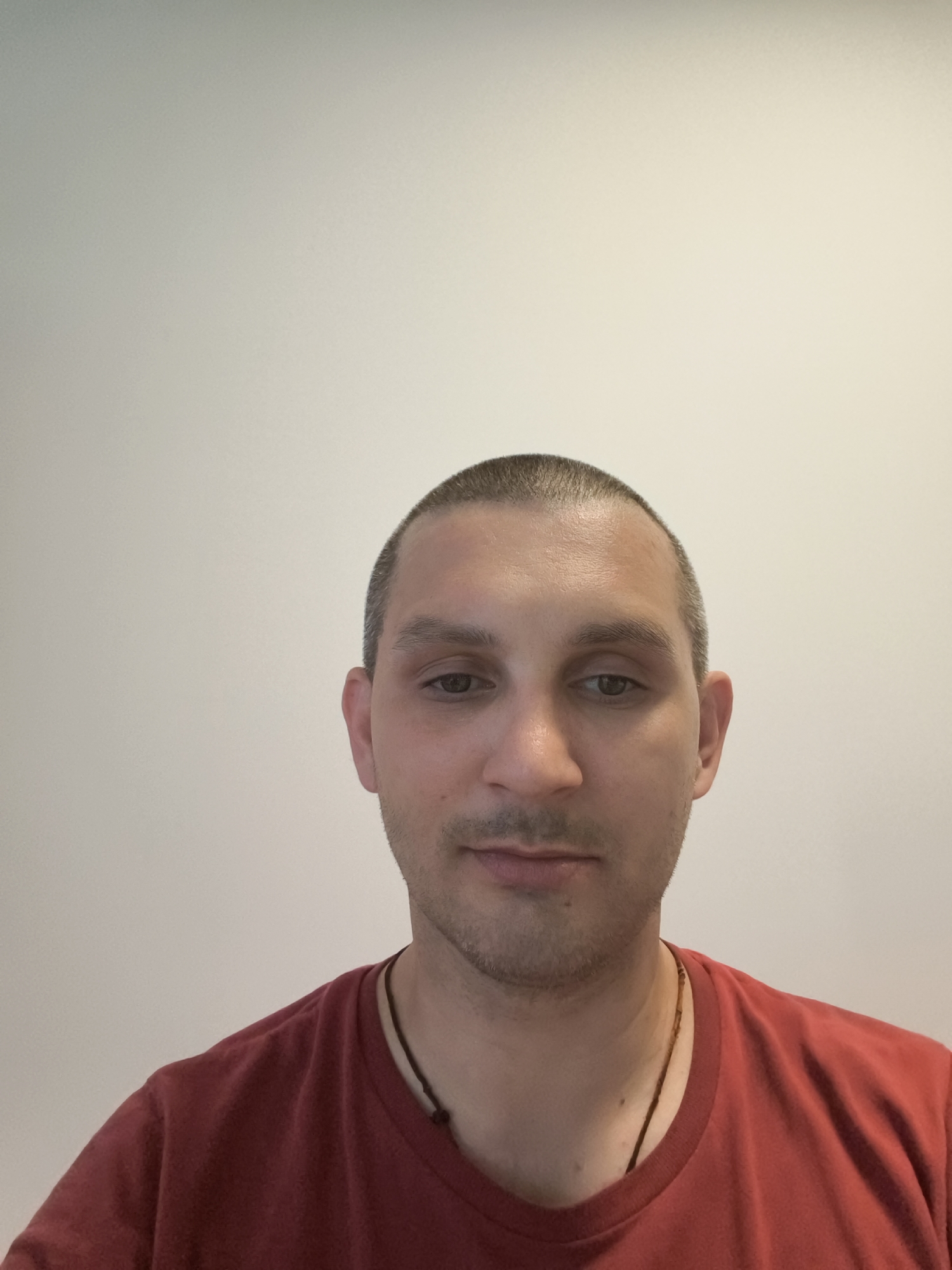 The image shows a medium shot of a man with a buzz cut, looking directly at the camera. He is wearing a red t-shirt and a dark necklace. The background is a plain, light-colored wall. There is no discernible information in the image that would indicate a specific location in the city of Wolgast, Germany. The image is a portrait with no contextual details about the environment or activities.