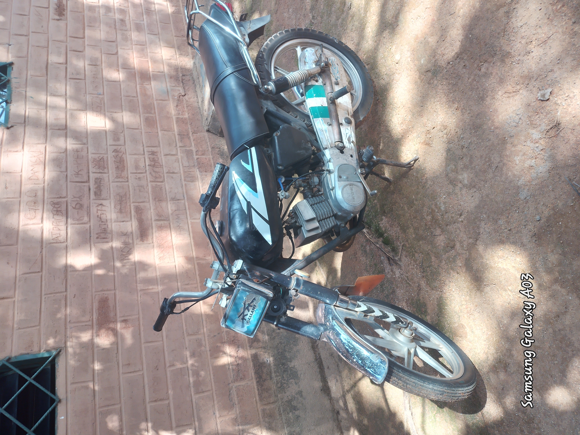 This is a medium-shot, eye-level, still photograph of a black motorcycle parked on a dirt surface in front of a brick wall. The motorcycle is positioned at a slight angle, with the front facing towards the viewer's left. It appears to be a Yamaha, based on the logo visible on the engine casing, and has a predominantly black exterior with white and blue graphics on the fuel tank. Chrome accents are present on the front fender, handlebars, and exhaust. The brick wall behind the motorcycle is textured and shows some graffiti or markings. A barred window is visible in the upper left corner of the frame. Sunlight casts shadows on the ground, suggesting it is daytime. The bottom right corner of the image displays the watermark "Samsung Galaxy A03". The scene is consistent with an outdoor setting in a residential or semi-urban area, possibly in Jos, Nigeria, given the style of the brickwork and the overall ambiance. There are no people visible in the photograph.