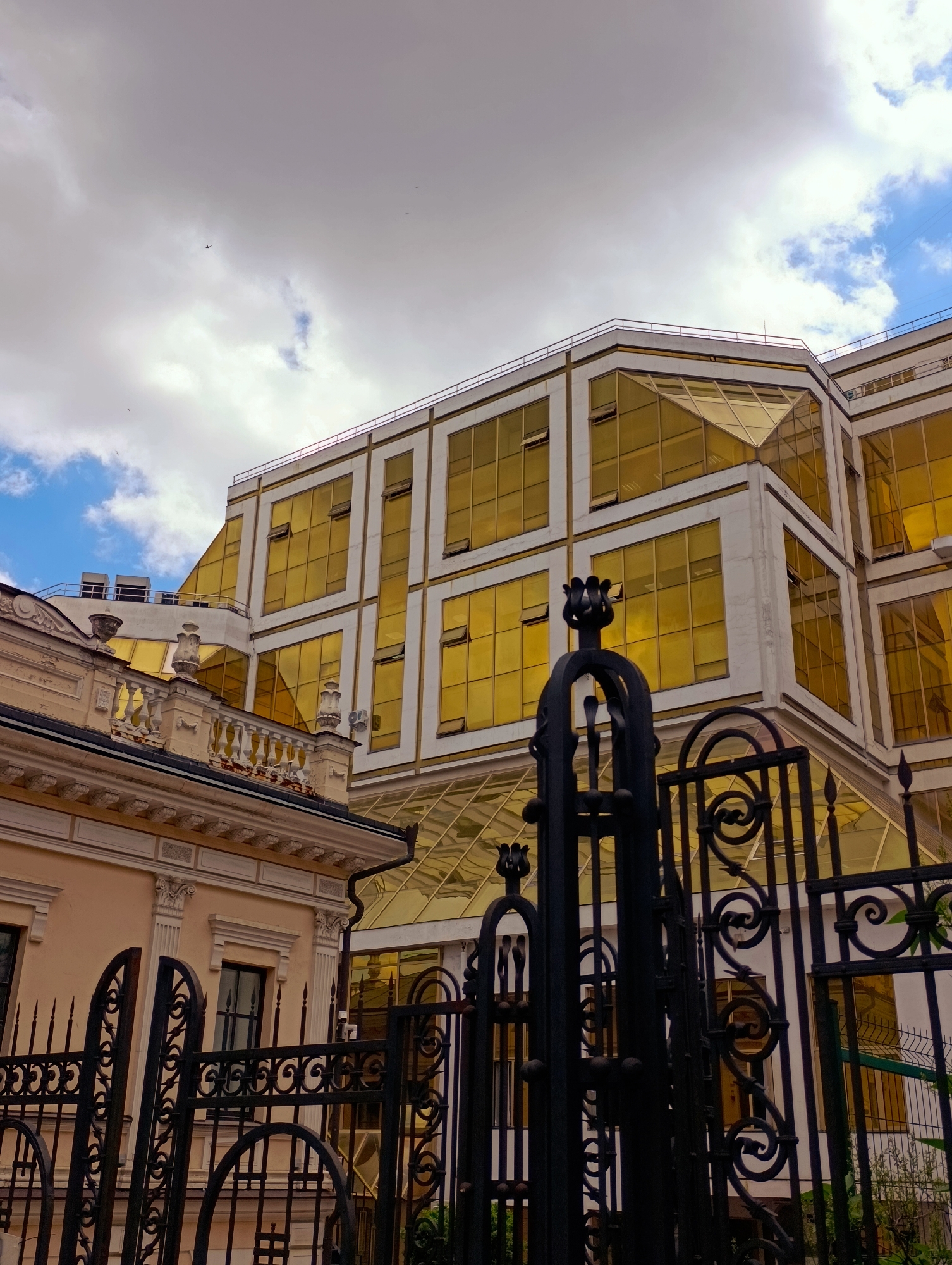 The image captures a view of the National Bank of Ukraine building in Kyiv, Ukraine. In the foreground, a black ornate metal gate with intricate scrollwork and finials partially obstructs the view. Behind the gate, an older, light-colored building with classical architectural details, including a balustrade and decorative cornices, is visible. To the right and behind this older building, the modern National Bank of Ukraine structure rises, characterized by its angular design and extensive use of golden-tinted glass windows. The sky above is a mix of white clouds and patches of blue, with a few distant birds or possibly airplanes visible. The scene depicts a juxtaposition of architectural styles within an urban setting.