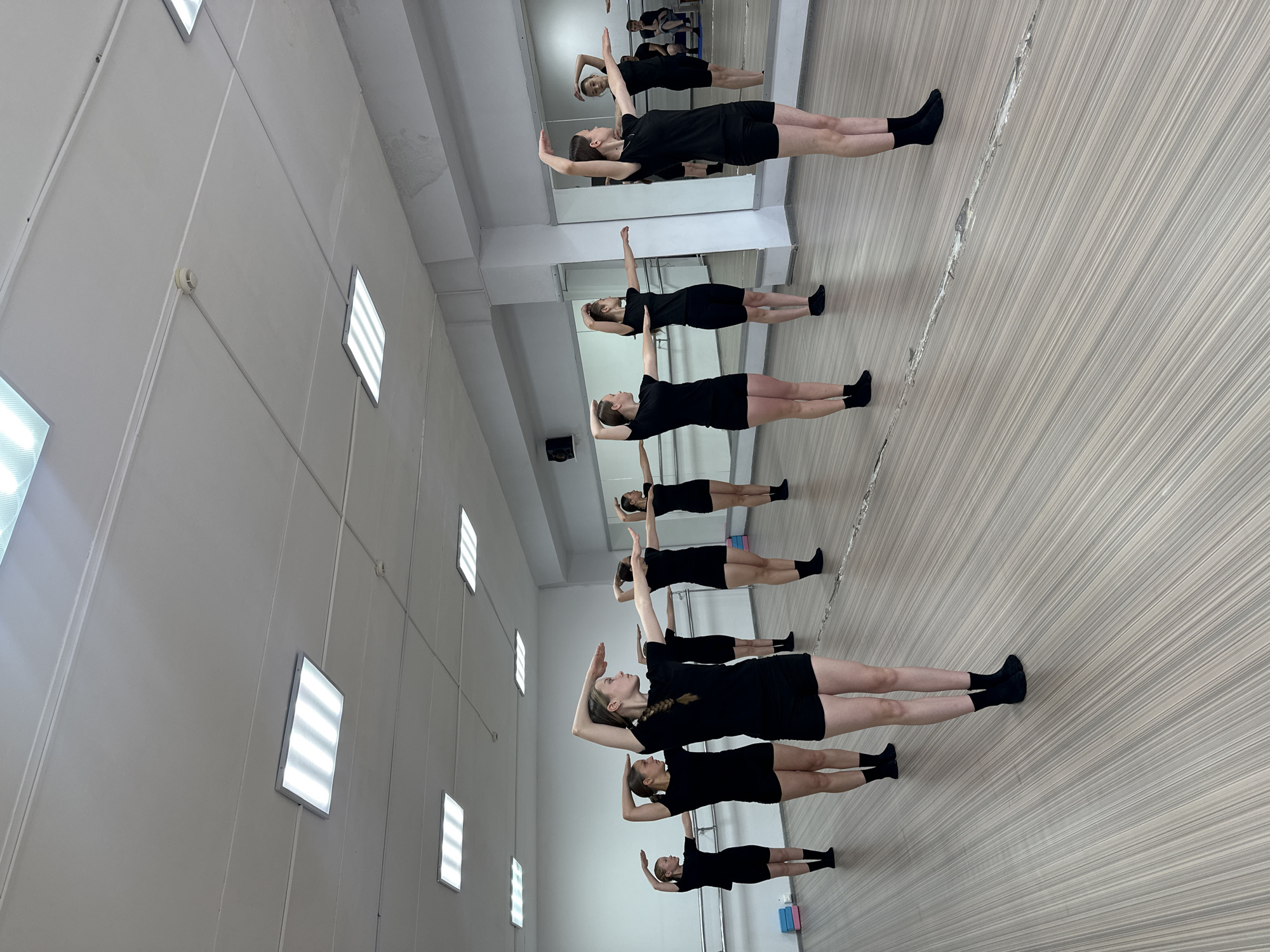 The image depicts a dance studio in Votkinsk, Russia, where a group of young women are practicing a choreographed routine. They are all dressed in black t-shirts and black shorts, with some wearing black socks. Their stances and arm positions suggest they are following instructions from an unseen instructor, possibly mirroring movements in the large mirrors that line the studio walls. The studio itself appears well-lit with fluorescent ceiling lights and has a polished wooden floor. A ballet barre is visible along the far wall, and stacked mats are positioned near the entrance. The overall atmosphere is one of focused practice and discipline within a professional dance environment.