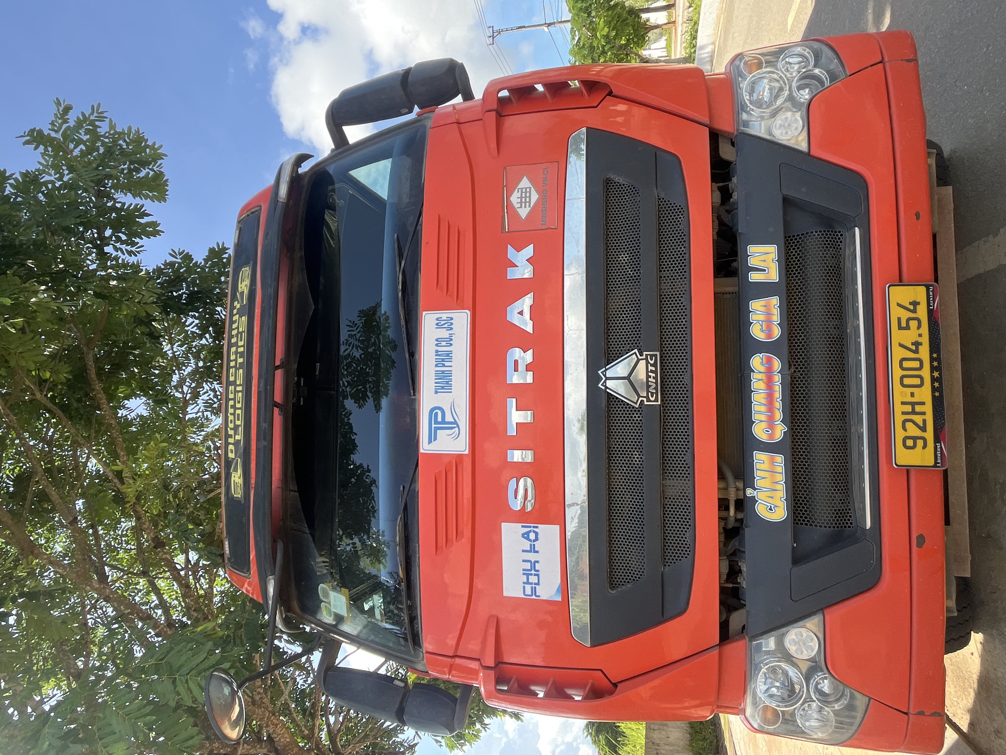 A front-view, medium shot captures a bright red SITRAK truck parked outdoors, possibly in Bo Y, Vietnam, under a tree with green foliage and a clear blue sky dotted with clouds. The truck's grille, featuring the "CNHTC" logo, is prominent. Above the grille, two rectangular stickers are visible: one with the text "TP THANH PHAT CO., JSC" and another with Vietnamese text and a red graphic. The truck's license plate reads "92H-004.54". The overall scene suggests a roadside setting with a focus on the vehicle.