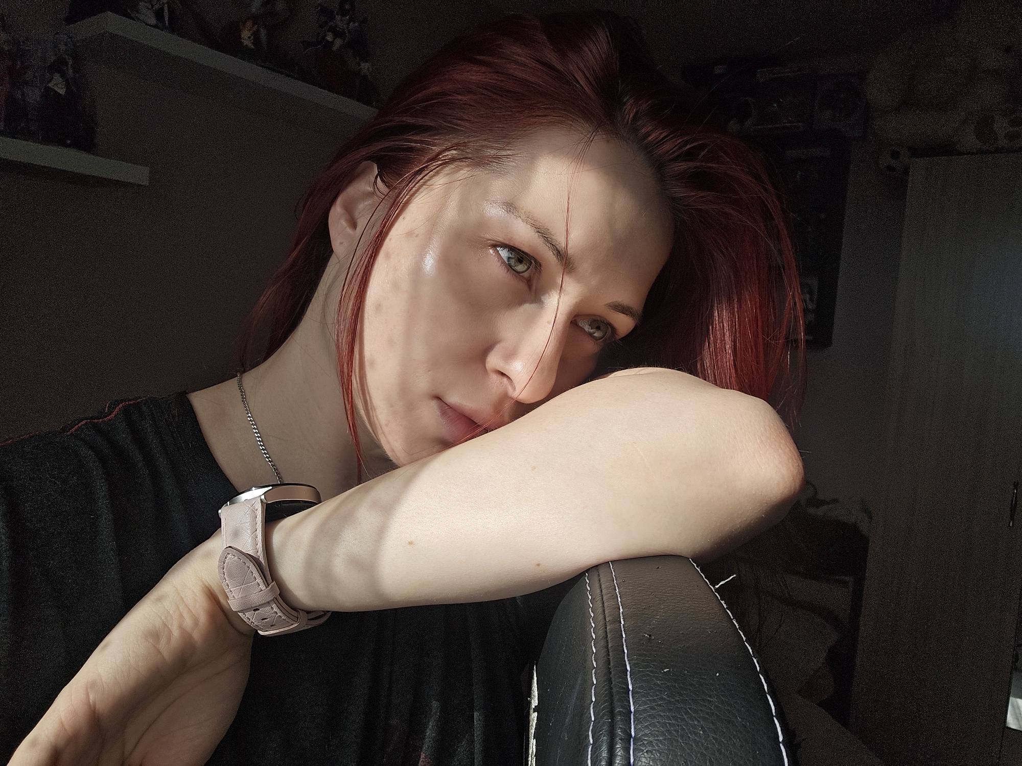 A person with red hair rests their head on their arm. They are wearing a pink watch and a silver necklace. The background includes a shelf with items and a dark cabinet. The scene is indoors with sunlight. No information indicates that this image was taken in Burgas, Bulgaria.