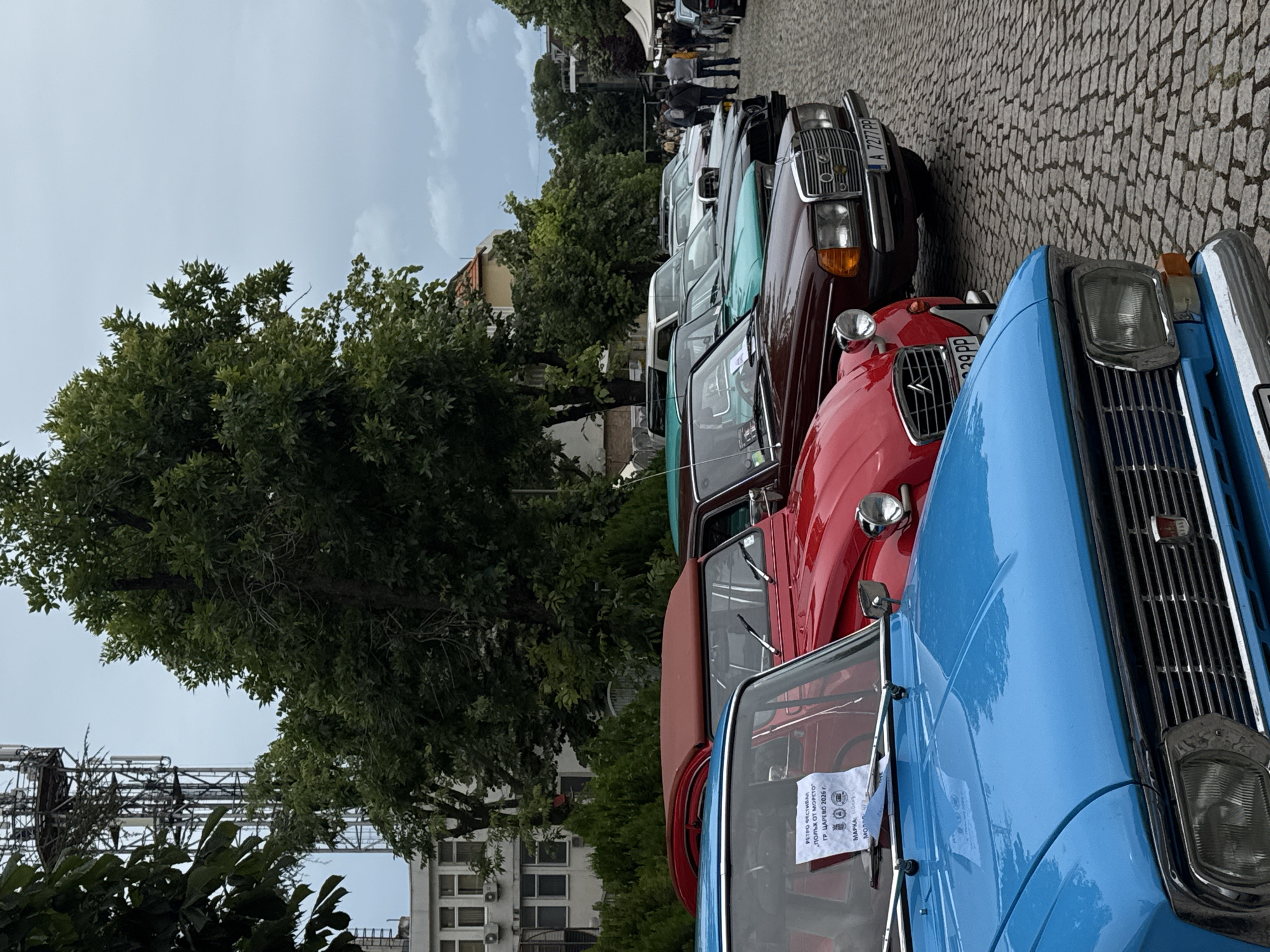 In Tsarevo, Bulgaria, a collection of vintage automobiles is displayed on a cobblestone street. The cars are parked in a row, facing right. In the foreground, a bright blue classic car is visible, followed by a red convertible, a dark maroon sedan, a teal sedan, and several other vehicles further down the line. The red car has a white sign on its windshield that reads "РЕТРО ФЕСТИВАЛ 'ПОЛЬК ОТ МОРЕТО' ГР. ЦАРЕВО 2026 г.". The background features mature trees, a building with several windows, and a tall telecommunications tower. A few people are scattered throughout the scene, some standing and observing the cars, others in the distance near tents. The sky is overcast, suggesting a cloudy day.