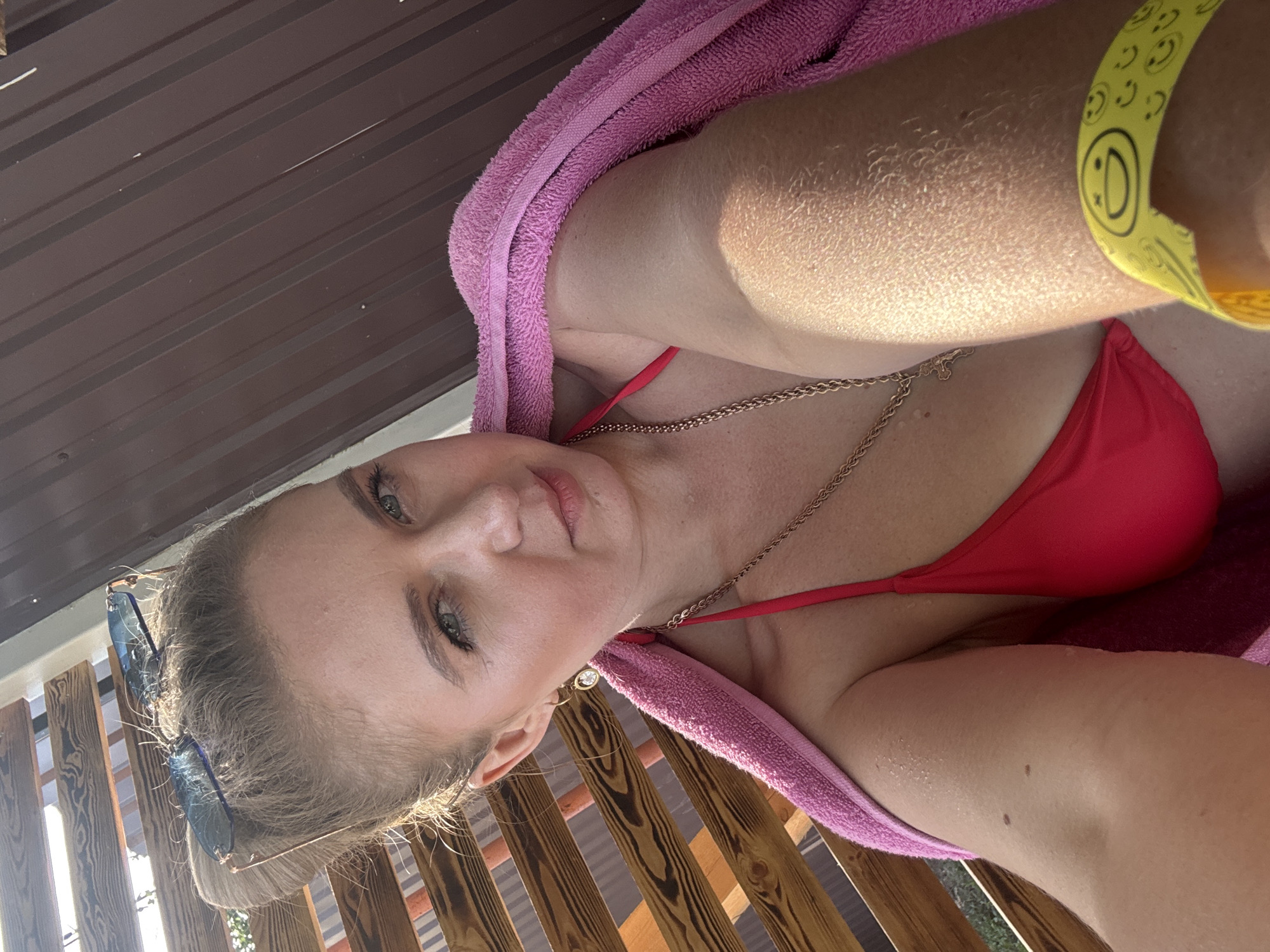 A woman with blonde hair pulled back in a bun, wearing sunglasses perched on her head, is pictured in a close-up selfie. She has on a red bikini top and a pink towel draped over her shoulder. A gold chain necklace with a cross pendant is visible around her neck, and she is wearing a hoop earring with a clear stone. Her right arm is extended, holding the camera, and a yellow wristband with smiley faces is on her wrist. The background consists of a wooden fence and a brown corrugated metal wall. The scene suggests an outdoor setting, possibly a poolside or beach area.