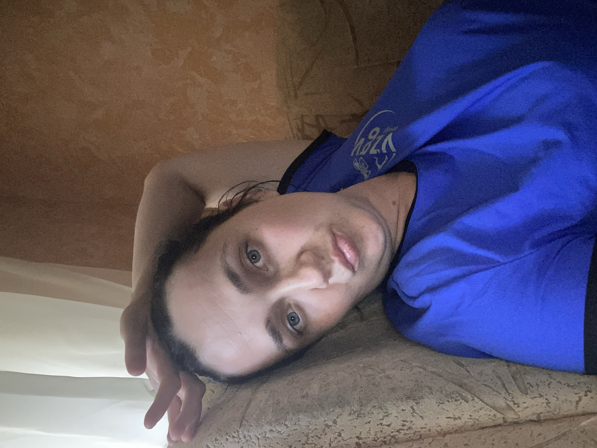 A woman is lying down on a couch with her right arm behind her head. She is wearing a blue t-shirt. She has light blue eyes and dark hair pulled back. The background consists of a beige couch, a window with white curtains to the left, and a textured orange wall to the right.