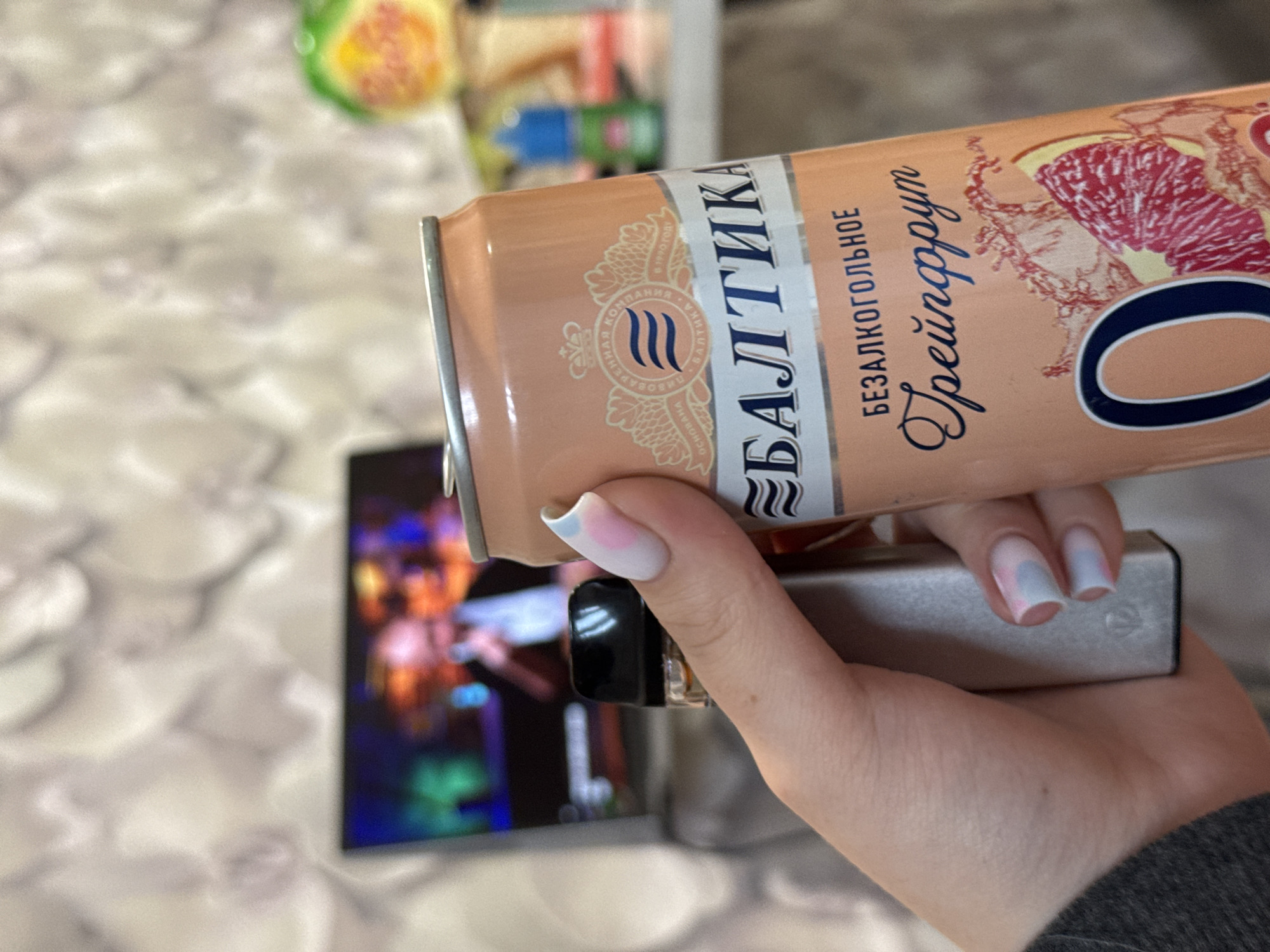 An indoor scene in Shirkovo, Russia, shows a person's hand holding two objects. The hand, with light skin and partially visible in a dark grey sleeve, has fingernails styled with a light base and soft pink and blue accents. In the left grip is a metallic grey rectangular device with a black top, likely a vape, featuring a subtle "V" logo near its base. To the right, the hand firmly grasps an aluminum can of Baltika 0% Grapefruit non-alcoholic beer. The can is peach-orange, displaying the brand "БАЛТИКА" (BALTIKA), the text "БЕЗАЛКОГОЛЬНОЕ" (NON-ALCOHOLIC) and "Грейпфрут" (Grapefruit), along with a large "O" and a graphic of a grapefruit slice with splashes. The can's upper section features a crowned emblem indicating "BREWING COMPANY BALTIKA" and "FOUNDED IN 1990." In the blurred background, a television screen displays a vibrant, colorful performance with a faint male figure holding a microphone. The wall behind is covered with a light-toned, subtly patterned wallpaper. To the right of the screen, out of focus, are a green and yellow object and a small blue bottle with a green cap on a surface.