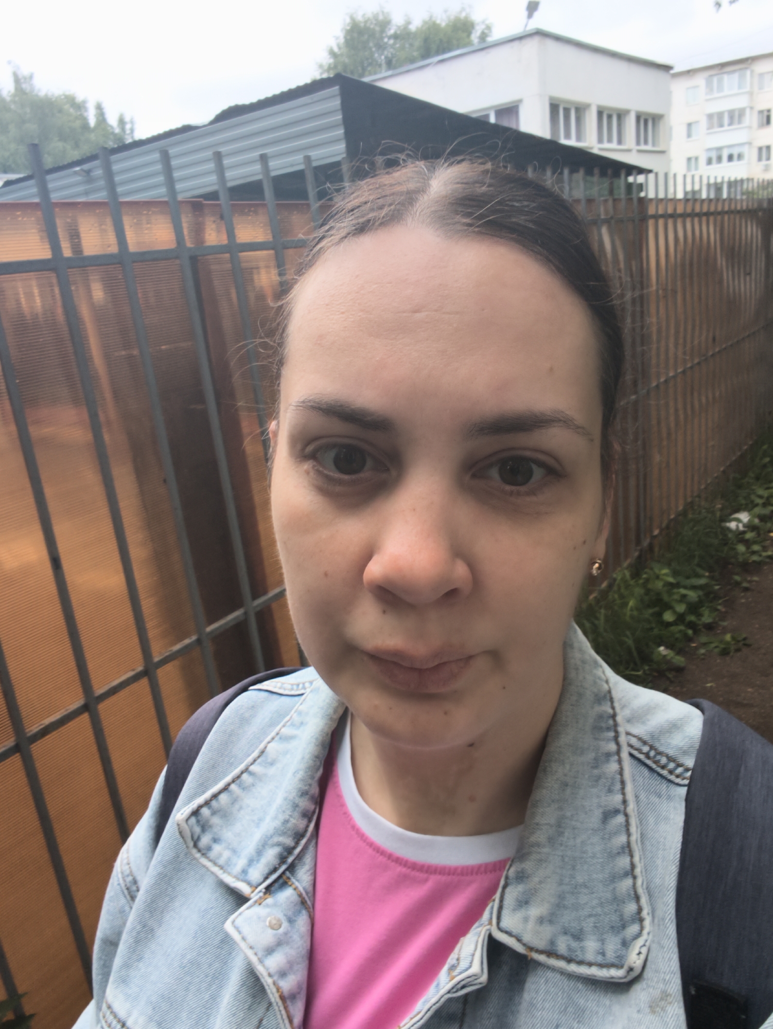 A frontal, waist-up selfie depicts an adult female outdoors in Chaikovsky, Russia, under an overcast sky. The subject has dark brown hair pulled back, displaying some visible flyaways around her forehead, and a neutral, direct gaze towards the camera. Her skin is fair, with minor blemishes and visible pores, particularly on her cheeks and under her eyes. She wears a faded light blue denim jacket over a bright pink t-shirt with a white collar trim. A dark-colored backpack strap is visible over her right shoulder. A small gold-colored stud earring is present in her right earlobe.
Behind her, a tall fence with dark vertical metal bars and translucent, corrugated orange-brown plastic panels runs across the midground. To the viewer's left, beyond the fence, dense green foliage and a dark-roofed shed-like structure are visible, topped by the overcast sky. To the viewer's right, the fence continues, bordering a dirt path with sparse green vegetation and some white litter. In the distant background are two multi-story light-colored residential buildings with numerous rectangular windows, characteristic of apartment complexes.