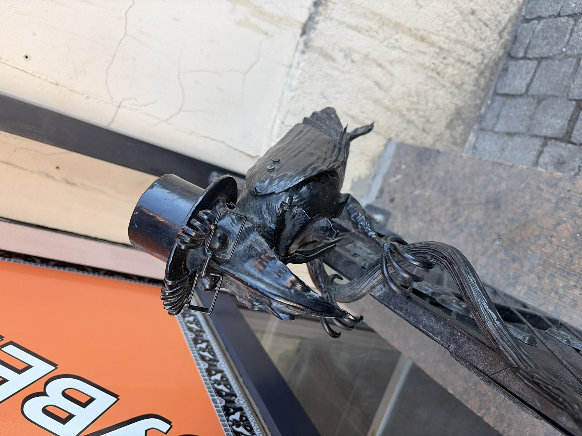 The image displays an angled, close-up view of an ornate, dark metal sculpture, likely an architectural embellishment or part of a gate. The primary subject is a stylized, grotesque figure resembling a bird or mythical creature, crafted from black metal and notably wearing a polished black top hat. The sculpture features intricate details suggesting feathers or scales, and its form is integrated with decorative metal scrollwork.
In the upper background, a light-colored building facade with visible cracks indicates an older structure. To the right, grey cobblestone paving is partially visible, hinting at an urban, historical setting. Below the metalwork, an orange sign with large white capital letters shows the partial text "BE", suggesting a commercial establishment or storefront. The scene is brightly illuminated, indicating daytime. No individuals are visible. The elaborate metalwork and traditional building materials are consistent with the architectural style found in Saint Petersburg, Russia.